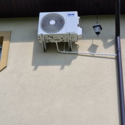 Klimator - Biała jednostka zewnętrzna klimatyzacji AUX zamontowana na beżowej elewacji budynku, widoczne rury odprowadzające skropliny i lampa zewnętrzna.