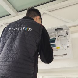 Klimator - Mężczyzna w firmowej kurtce 'KLIMAT' montuje klimatyzator na białej ścianie. Widok od tyłu, skupienie na urządzeniu i detalu kurtki.