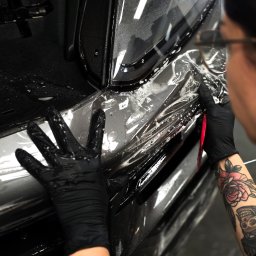 Jadeit Studio Beata Szyszko - Aplikacja folii ochronnej na błotnik czarnego auta. Widoczne dłonie w czarnych rękawiczkach, narzędzie i fragment tatuażu na ręce osoby aplikującej.