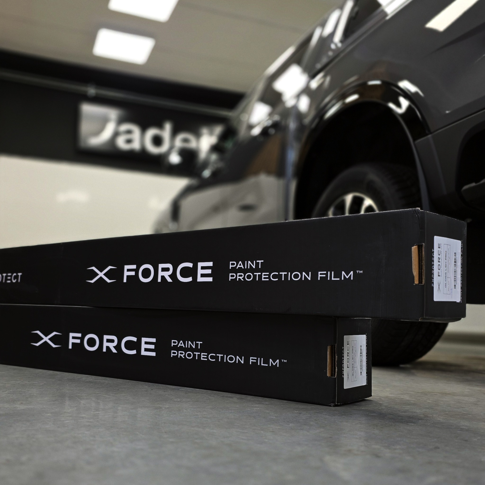 Dwa opakowania folii ochronnej X Force Paint Protection Film na tle czarnego SUV-a w warsztacie. Ostrość na opakowaniach, tło rozmyte.