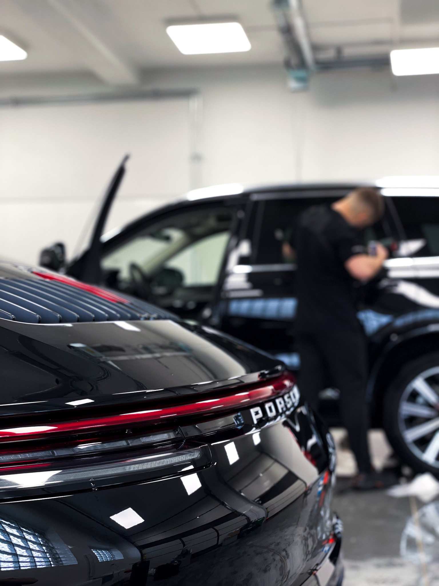 Błyszczący, czarny Porsche z czerwonym akcentem na tylnych światłach, w tle pracownik detailingu przy czarnym SUV-ie. Polerowanie lakieru w jasnym garażu.