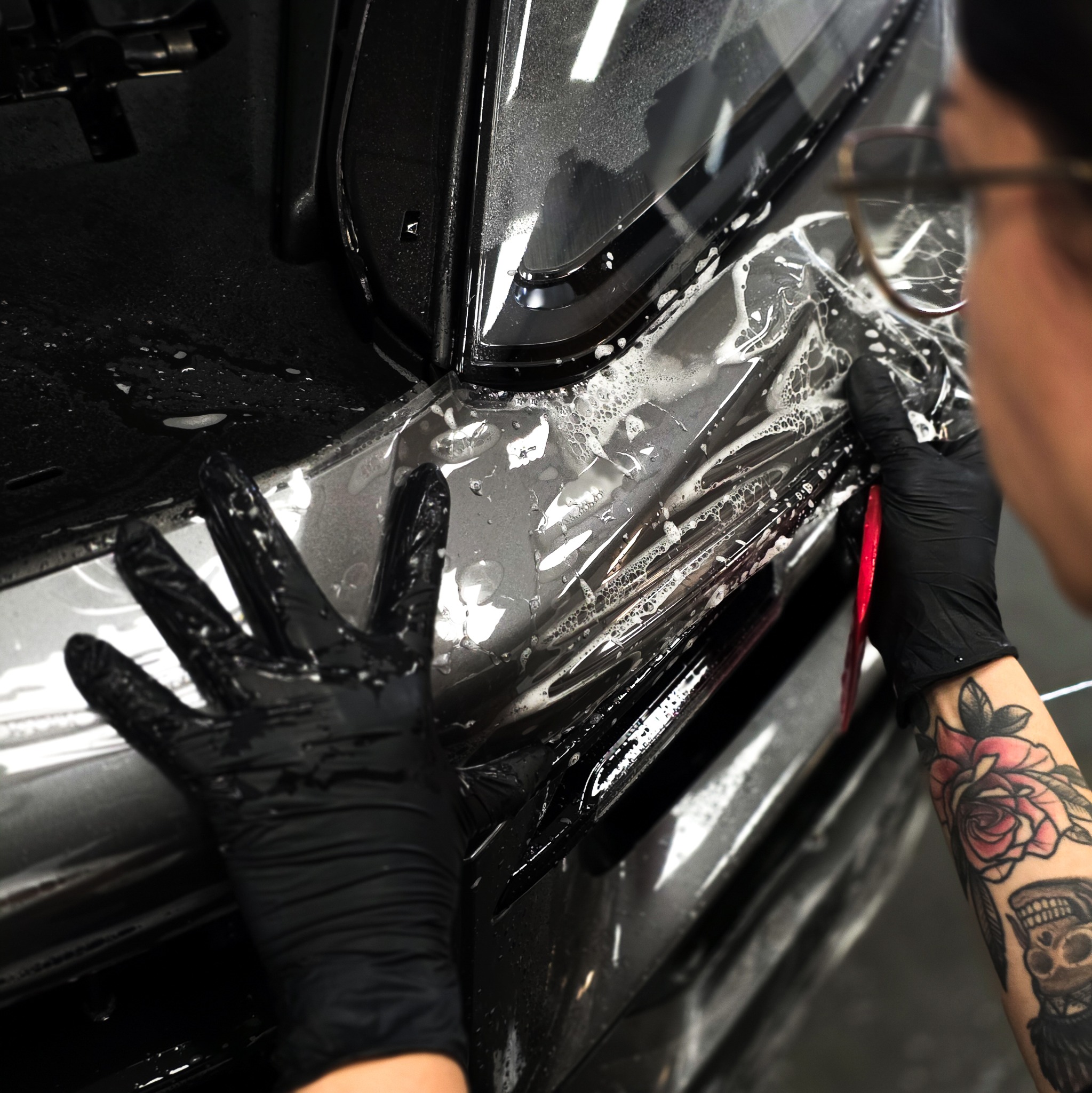 Aplikacja folii ochronnej na błotnik czarnego auta. Widoczne dłonie w czarnych rękawiczkach, narzędzie i fragment tatuażu na ręce osoby aplikującej.