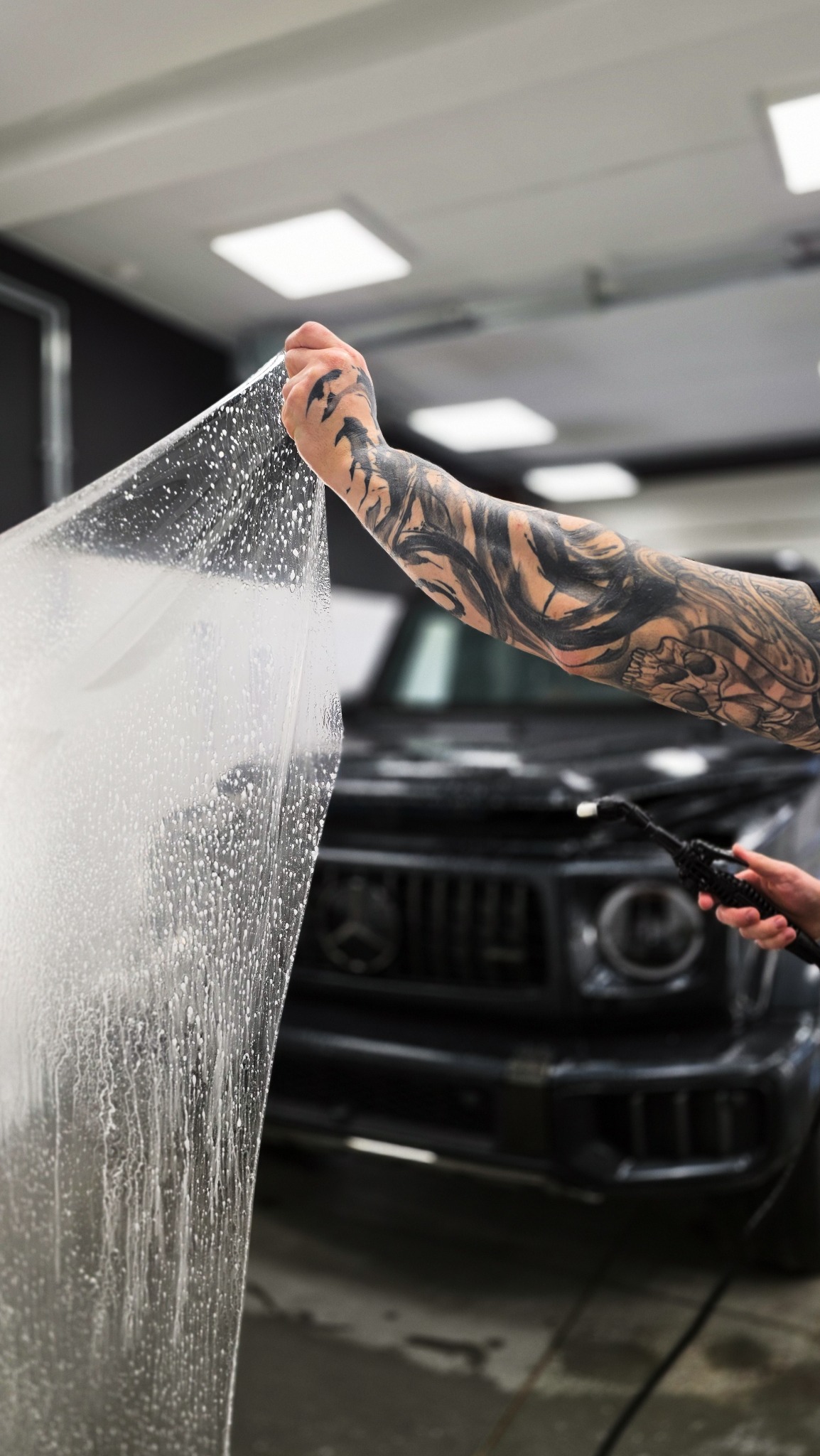 Aplikacja folii ochronnej na czarny samochód. Widoczna wytatuowana ręka pracownika i narzędzie do aplikacji. Bąbelki wody pod folią. Detailing auta.