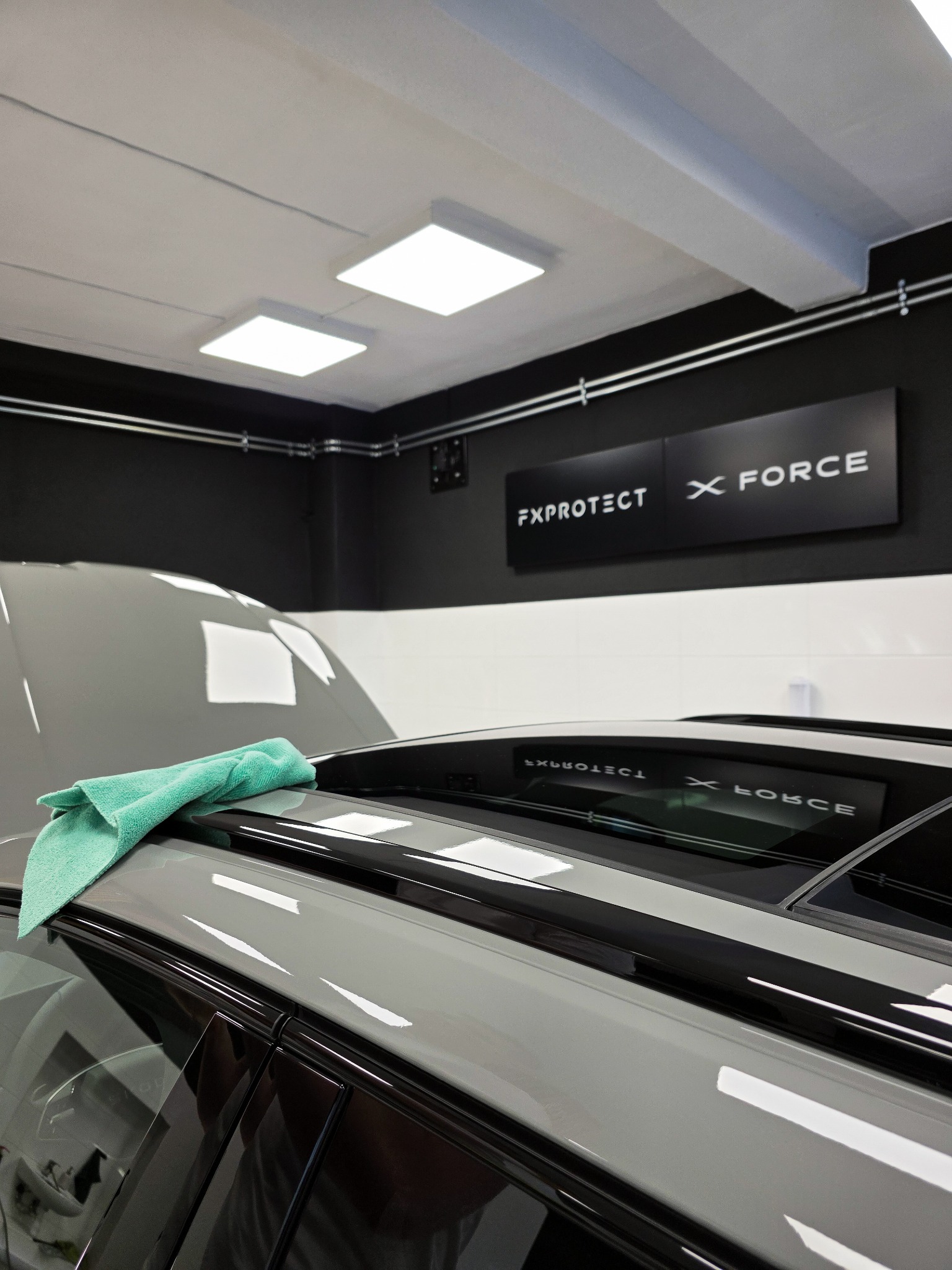 Błyszczący, szary samochód po detailingu, z zieloną ściereczką na dachu. Widoczne logo 'FXPROTECT X FORCE' na ścianie. Oświetlenie studyjne.