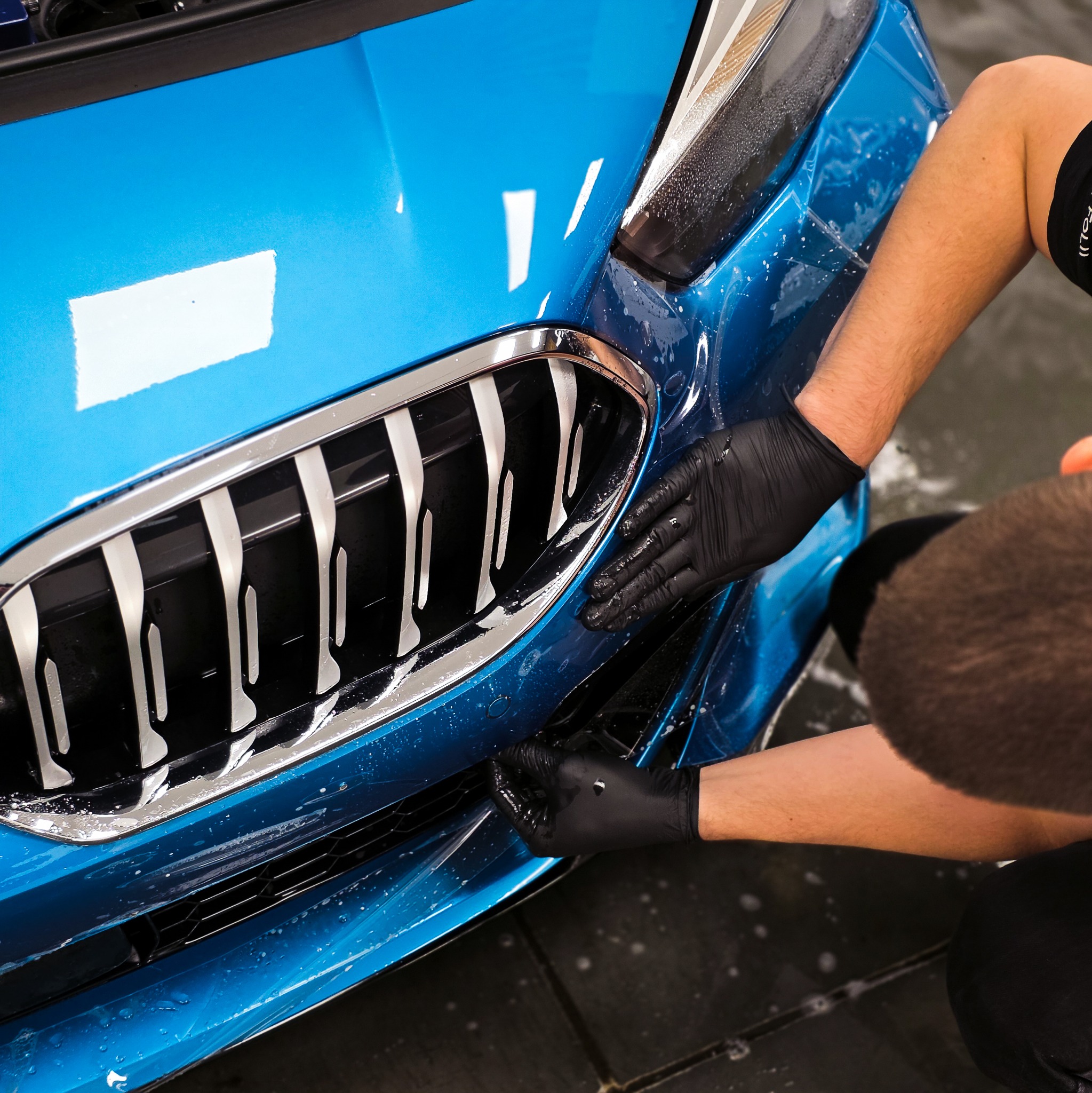 Aplikacja folii ochronnej na błękitny samochód. Widoczne dłonie w czarnych rękawicach, fragment grilla i przedniego zderzaka. Detailing auta.