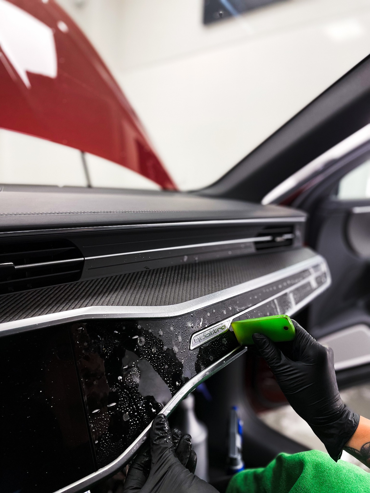 Aplikacja folii ochronnej na panel deski rozdzielczej Audi Quattro. Widoczne dłonie w czarnych rękawicach, narzędzie i płyn. Ochrona wnętrza pojazdu.