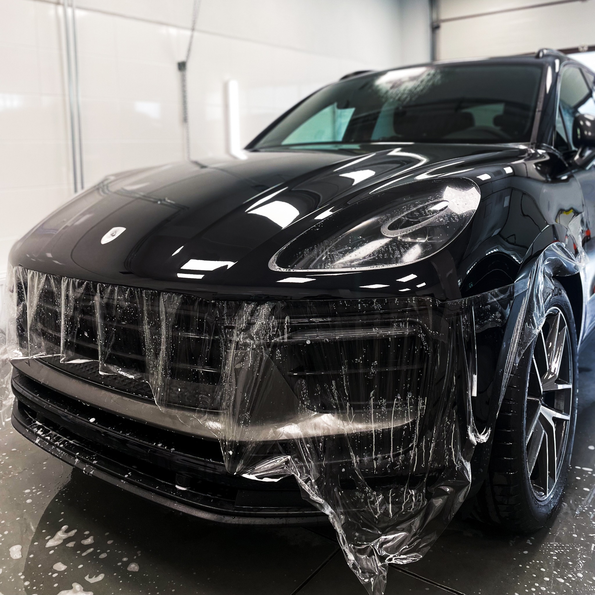 Czarny samochód Porsche, zabezpieczony folią ochronną na przedniej części, w trakcie aplikacji w myjni. Widoczne detale felg i reflektorów.