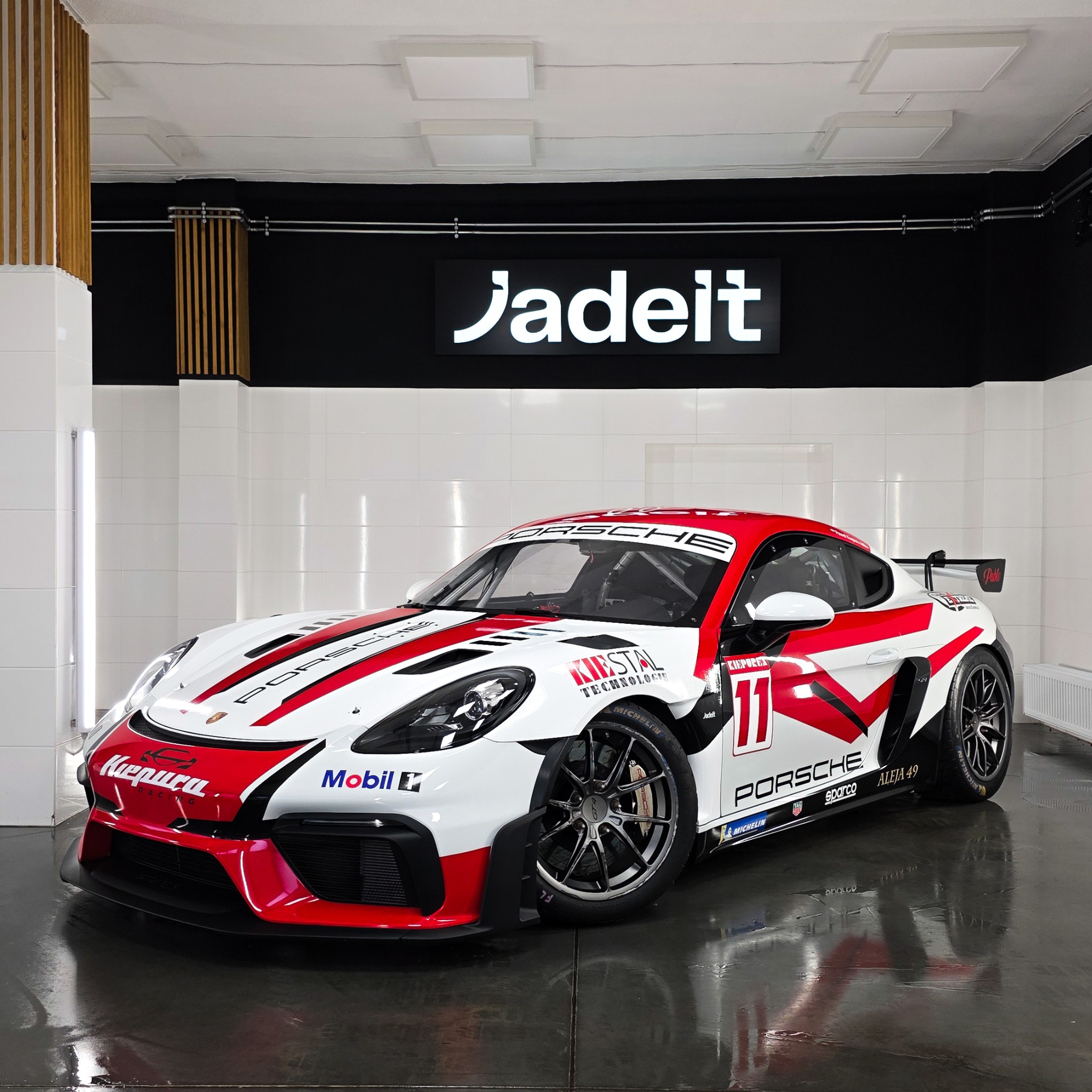 Porsche 718 Cayman GT4 Clubsport z czerwono-białym oklejeniem w sterylnym garażu z logo Jadeit. Błyszcząca podłoga odbija światło. Widoczne detale: numer 11, logotypy sponsorów.