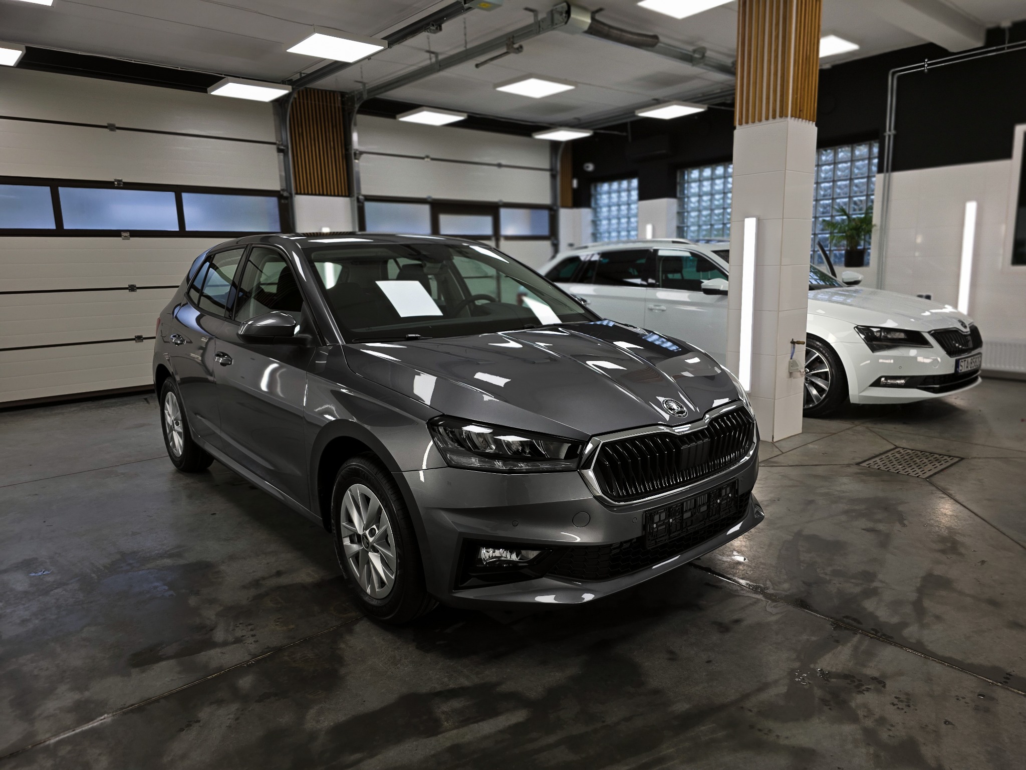 Szara Skoda Fabia w studiu detailingowym, widok z przodu. W tle biały samochód i brama garażowa. Błyszcząca podłoga, profesjonalne oświetlenie.