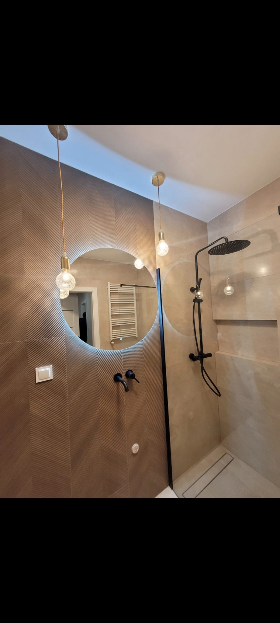 Nowoczesna łazienka z okrągłym lustrem LED, czarną armaturą i płytkami w jodełkę. Widoczny prysznic typu walk-in z odpływem liniowym i wiszące lampy z żarówkami Edisona.