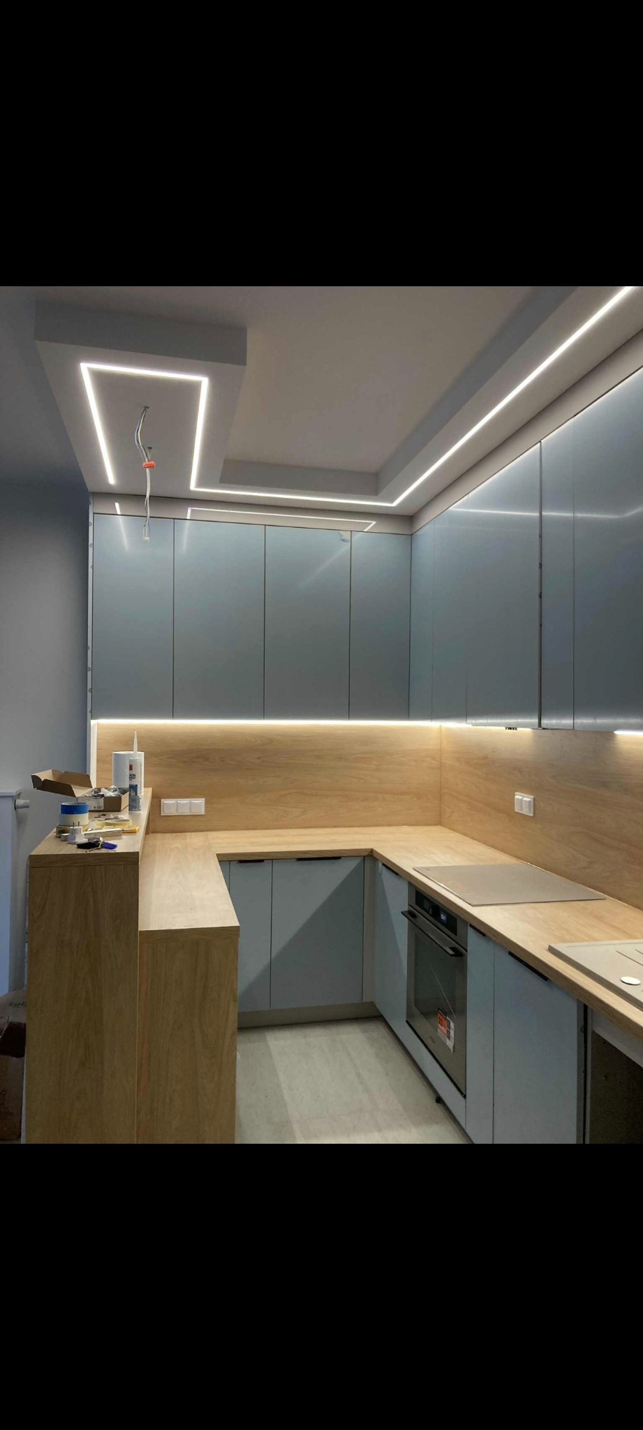 Nowoczesna kuchnia w odcieniach szarości i drewna z oświetleniem LED. Widoczne szafki, blat, piekarnik i elementy instalacji elektrycznej na suficie.