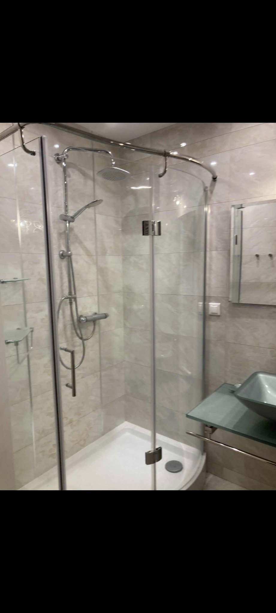 Nowoczesna kabina prysznicowa z deszczownicą i szklaną umywalką na tle beżowych płytek. Widoczne chromowane elementy i minimalistyczny design.