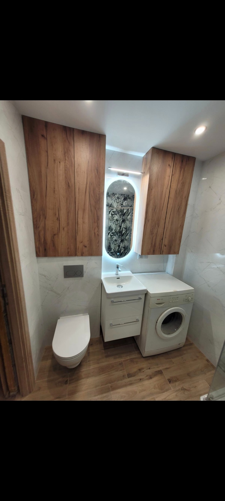 Nowoczesna łazienka z drewnianymi szafkami, owalnym lustrem z podświetleniem LED, białą umywalką z szafką i pralką. Podłoga wyłożona płytkami imitującymi drewno.