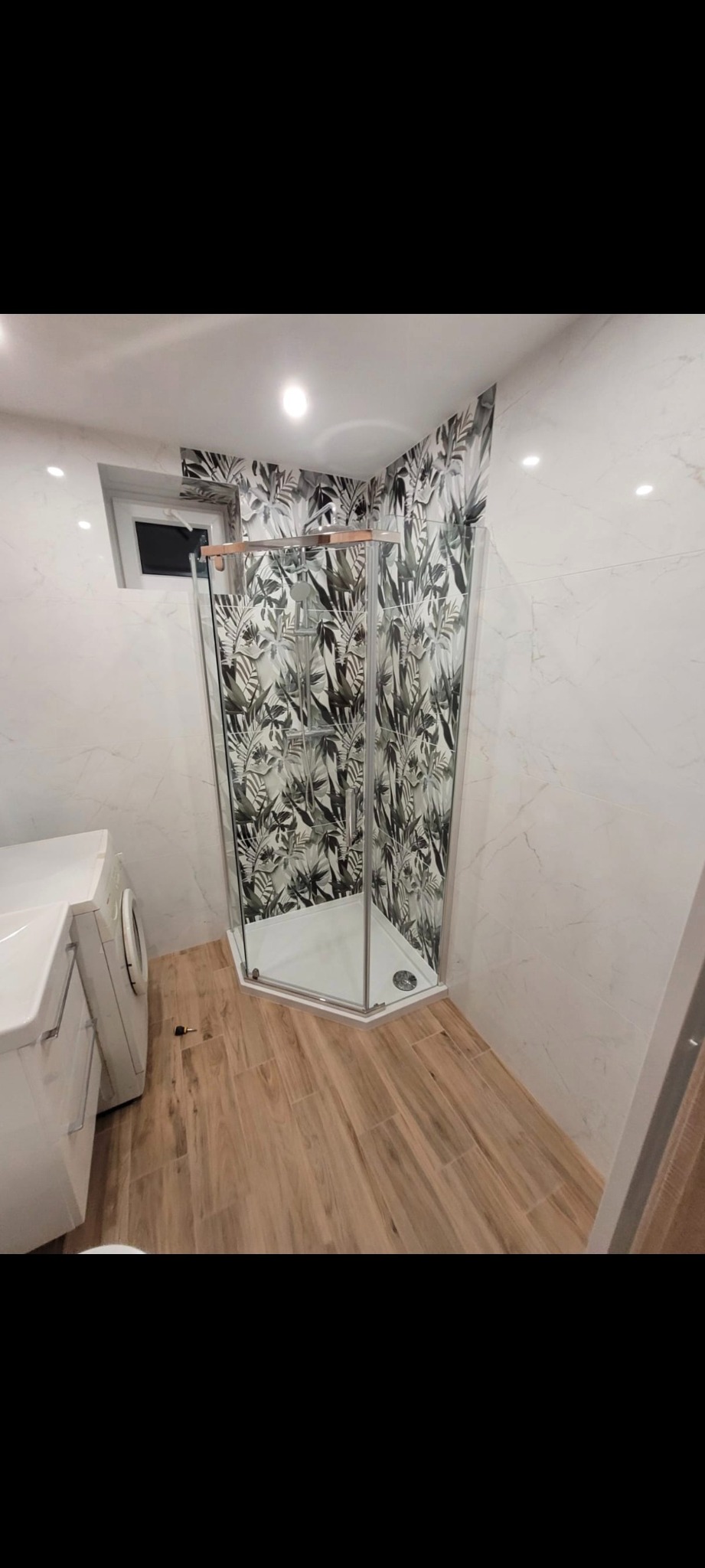 Nowoczesna łazienka po remoncie: kabina prysznicowa z motywem liści, drewnopodobne płytki podłogowe, marmurowe ściany i pralka. Jasne oświetlenie.