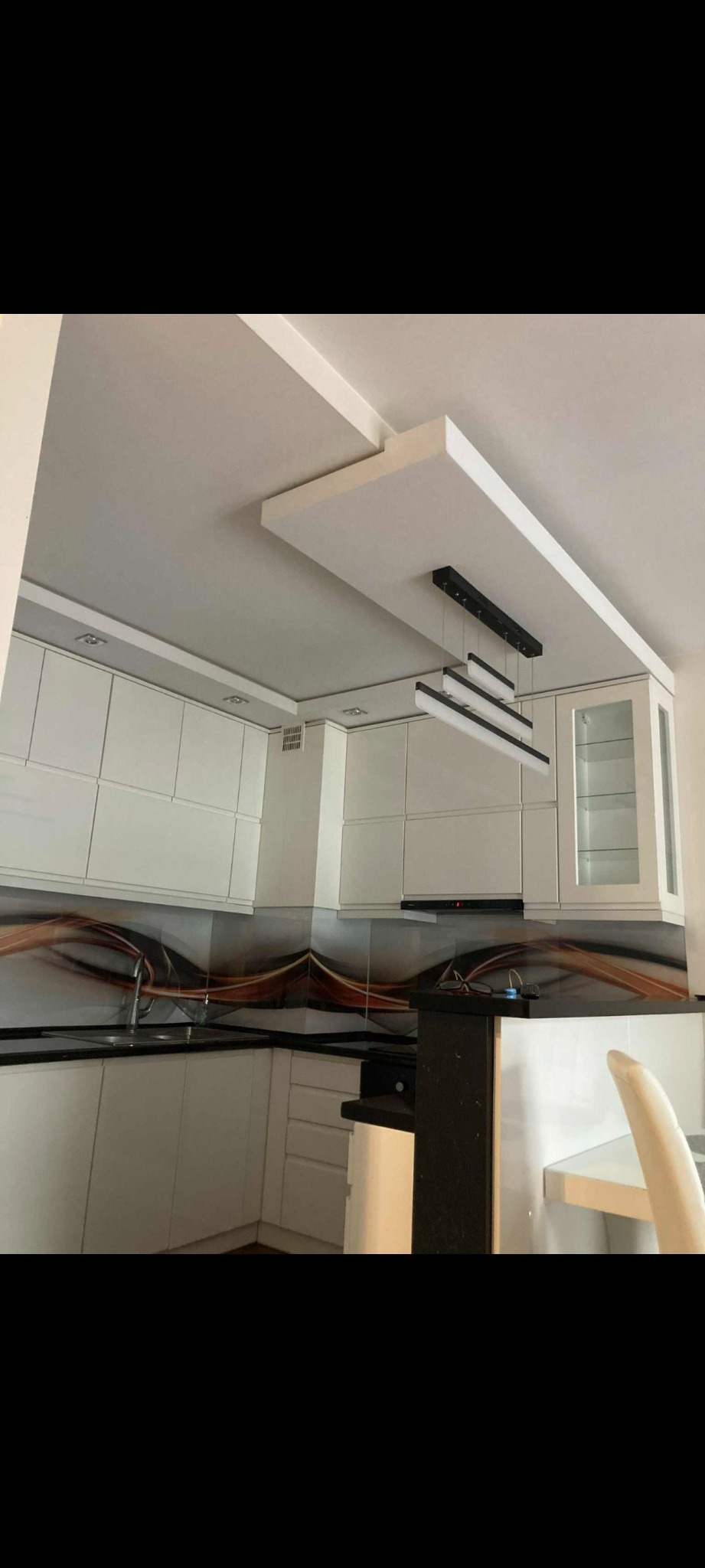 Nowoczesna kuchnia z białymi szafkami, czarnym blatem i dekoracyjnym panelem nad blatem. Podwieszany sufit z oświetleniem. Ujęcie z dołu, podkreślające przestrzeń.