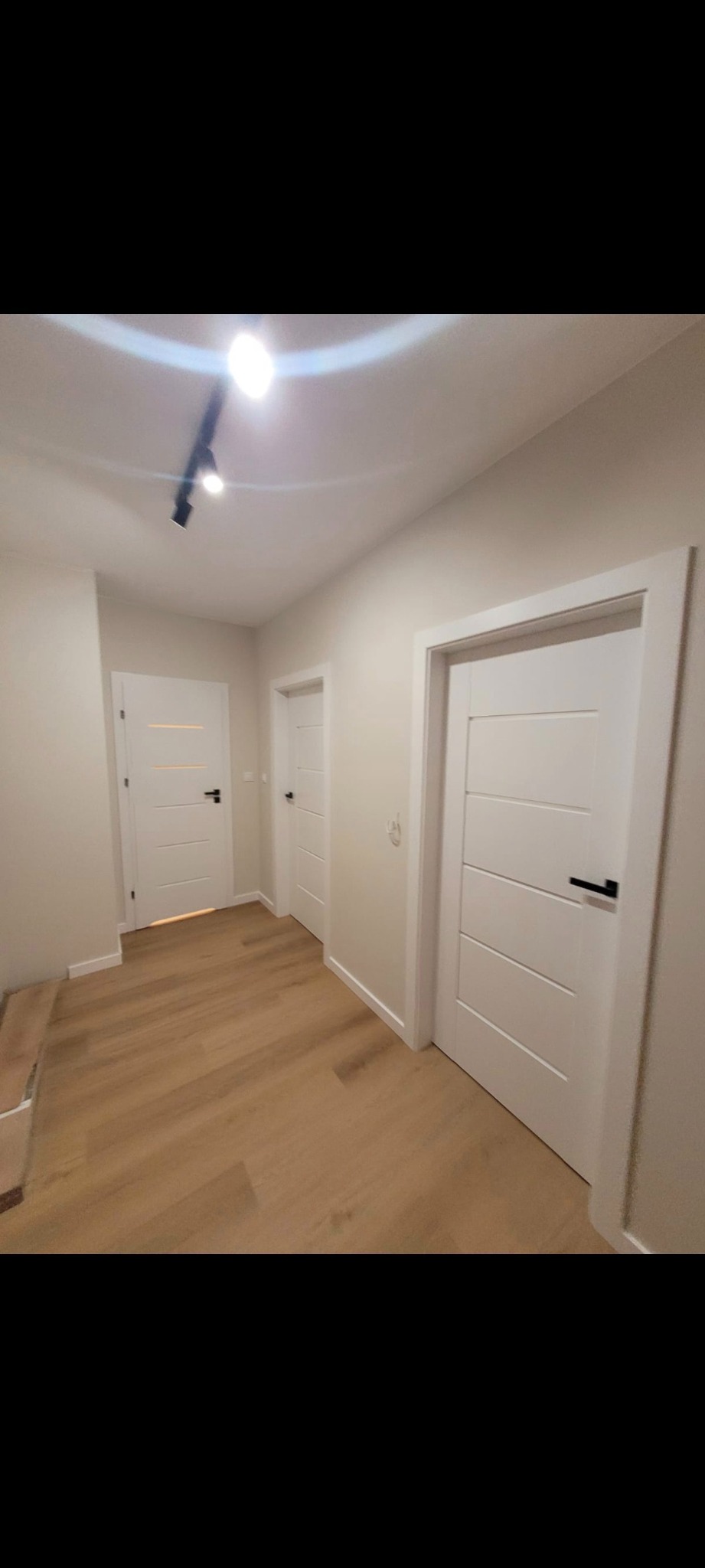 Jasny korytarz z trzema białymi drzwiami z czarnymi klamkami i panelami podłogowymi. Oświetlenie sufitowe punktowe. Minimalistyczny design.