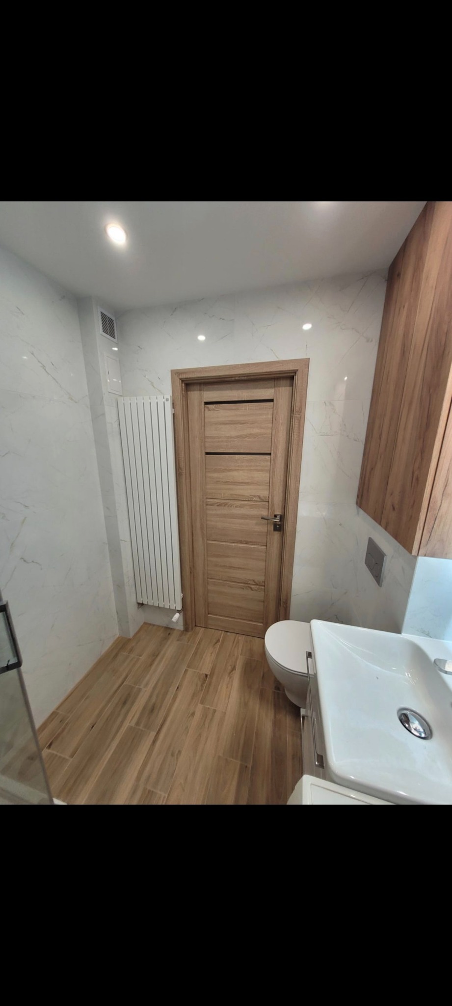 Nowoczesna łazienka po remoncie: drewnopodobne płytki, marmurowe ściany, drewniana szafka i biała armatura. Widok na drzwi i grzejnik.