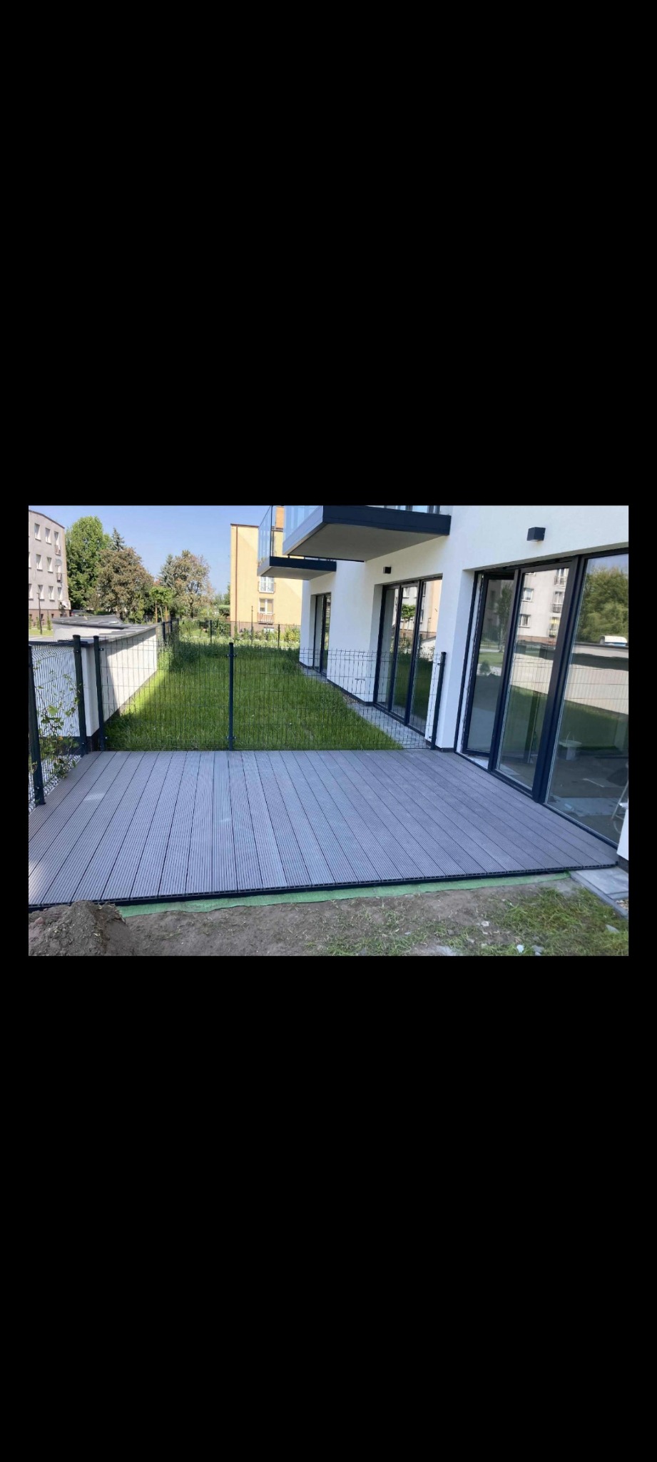 Nowoczesny taras z szarych desek kompozytowych przy budynku z dużymi oknami. Widok na trawnik i ogrodzenie. Słoneczny dzień.