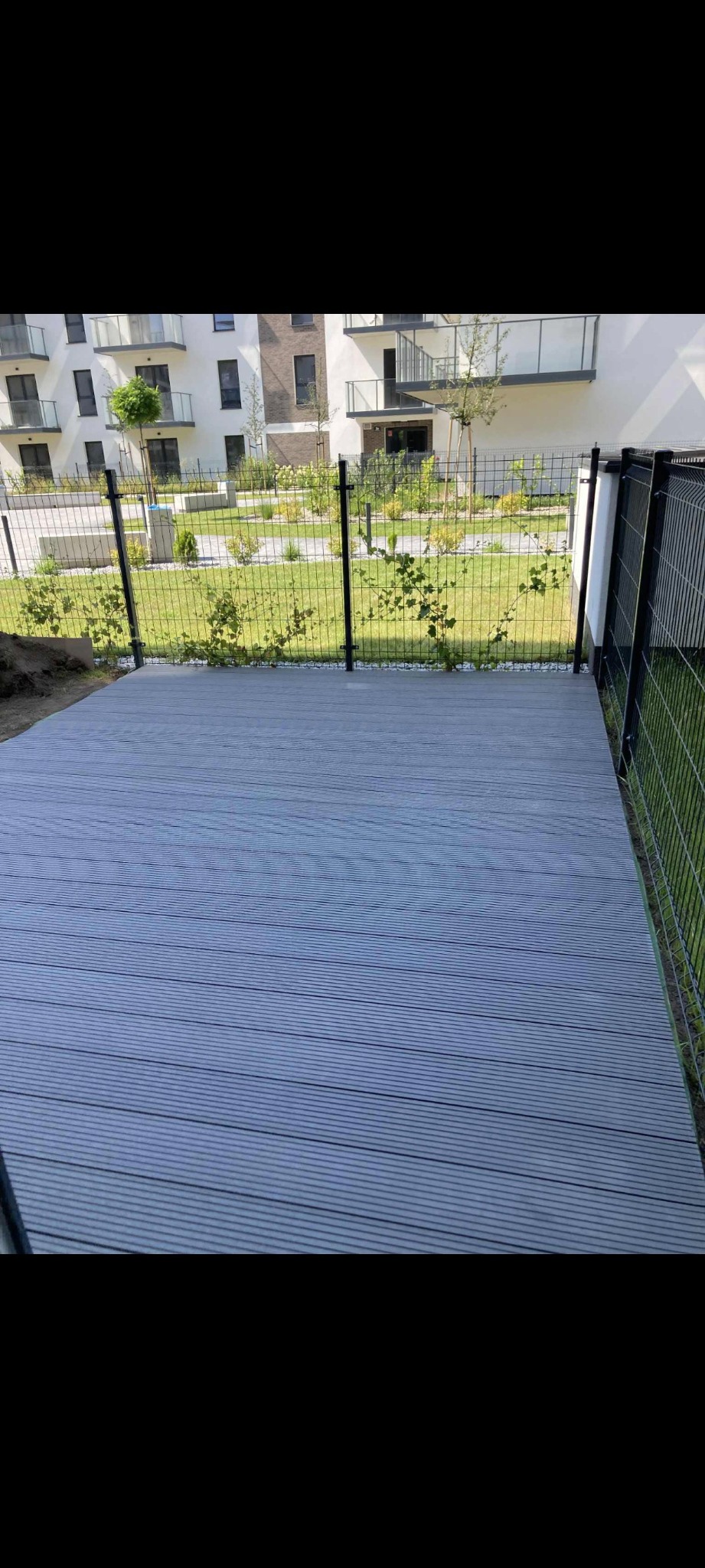 Szara deska kompozytowa tworząca taras przy ogrodzeniu na tle nowoczesnego budynku mieszkalnego z balkonami. Widok z poziomu gruntu.