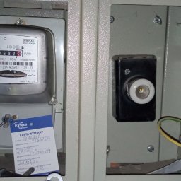 Wnętrze skrzynki elektrycznej z zamontowanym licznikiem energii, widoczna karta wymiany Enea Operator, przewody uziemiające i bezpiecznik. Szare tło obudowy.