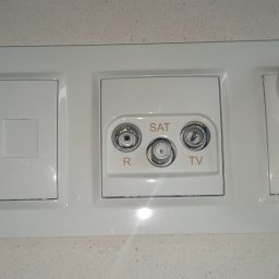 Biały panel z gniazdem RJ45, gniazdem antenowym SAT/TV i podwójnym gniazdkiem elektrycznym, zamontowany na jasnej ścianie. Widok z bliska, oświetlenie naturalne.