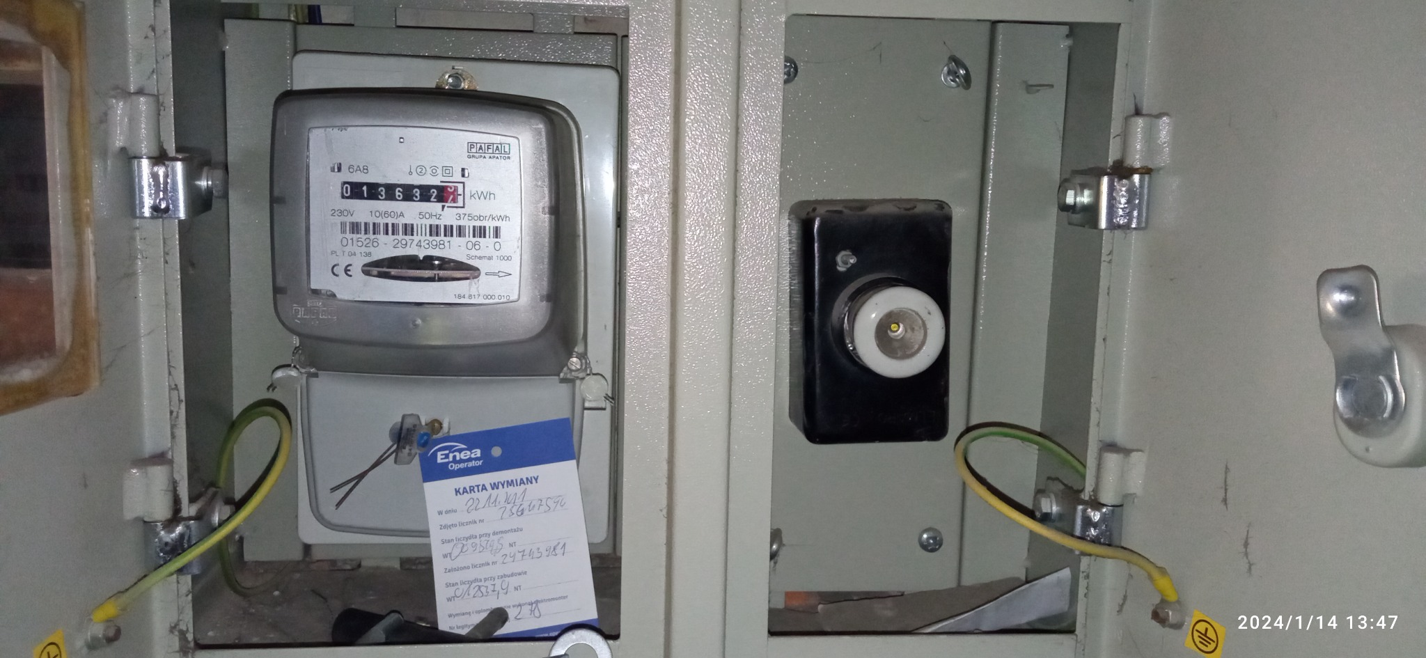 Wnętrze skrzynki elektrycznej z zamontowanym licznikiem energii, widoczna karta wymiany Enea Operator, przewody uziemiające i bezpiecznik. Szare tło obudowy.