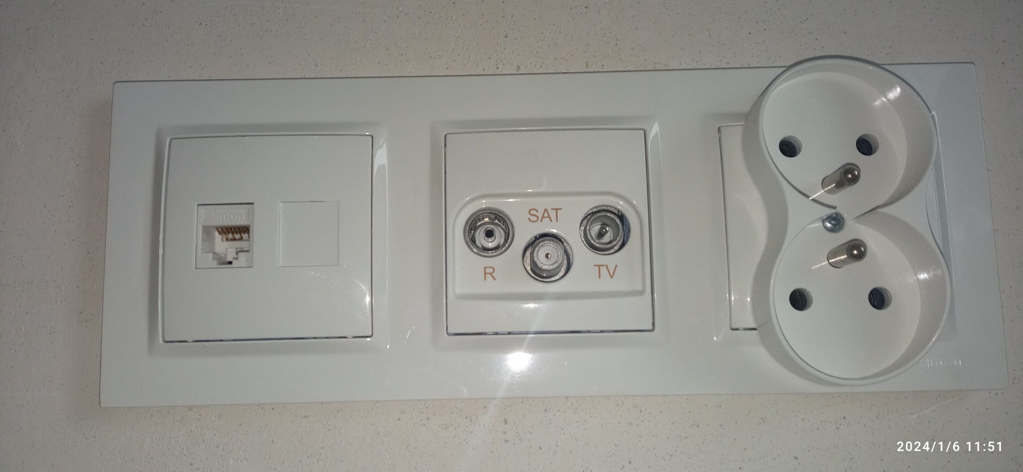 Biały panel z gniazdem RJ45, gniazdem antenowym SAT/TV i podwójnym gniazdkiem elektrycznym, zamontowany na jasnej ścianie. Widok z bliska, oświetlenie naturalne.