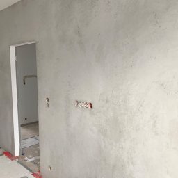 PHOTOPORT.TECH MACIEJ JAWORSKI - Szara ściana z efektem betonu w nowym wnętrzu, widoczne puszki elektryczne i fragment otwartych drzwi. Minimalistyczny design z surowym wykończeniem.