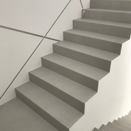 PHOTOPORT.TECH MACIEJ JAWORSKI - Minimalistyczne schody z szarego materiału, prawdopodobnie beton architektoniczny, z geometrycznymi detalami na ścianie. Widok z dołu, podkreślający prostotę i nowoczesny design.