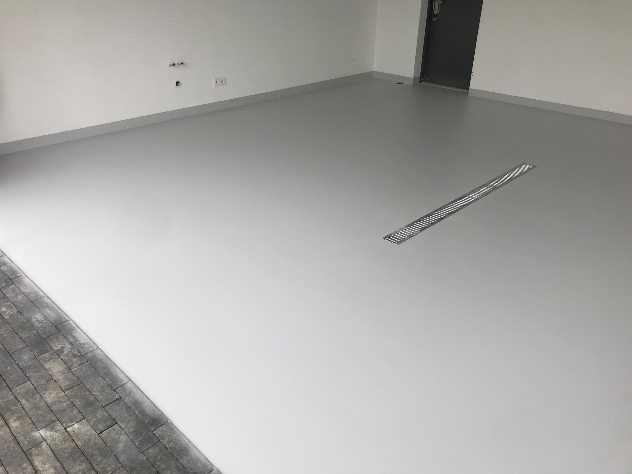 Jasnoszara, gładka posadzka w garażu z odpływem liniowym. Widoczne białe ściany i fragment kostki brukowej przy wjeździe. Minimalistyczny design, świeżo po realizacji.
