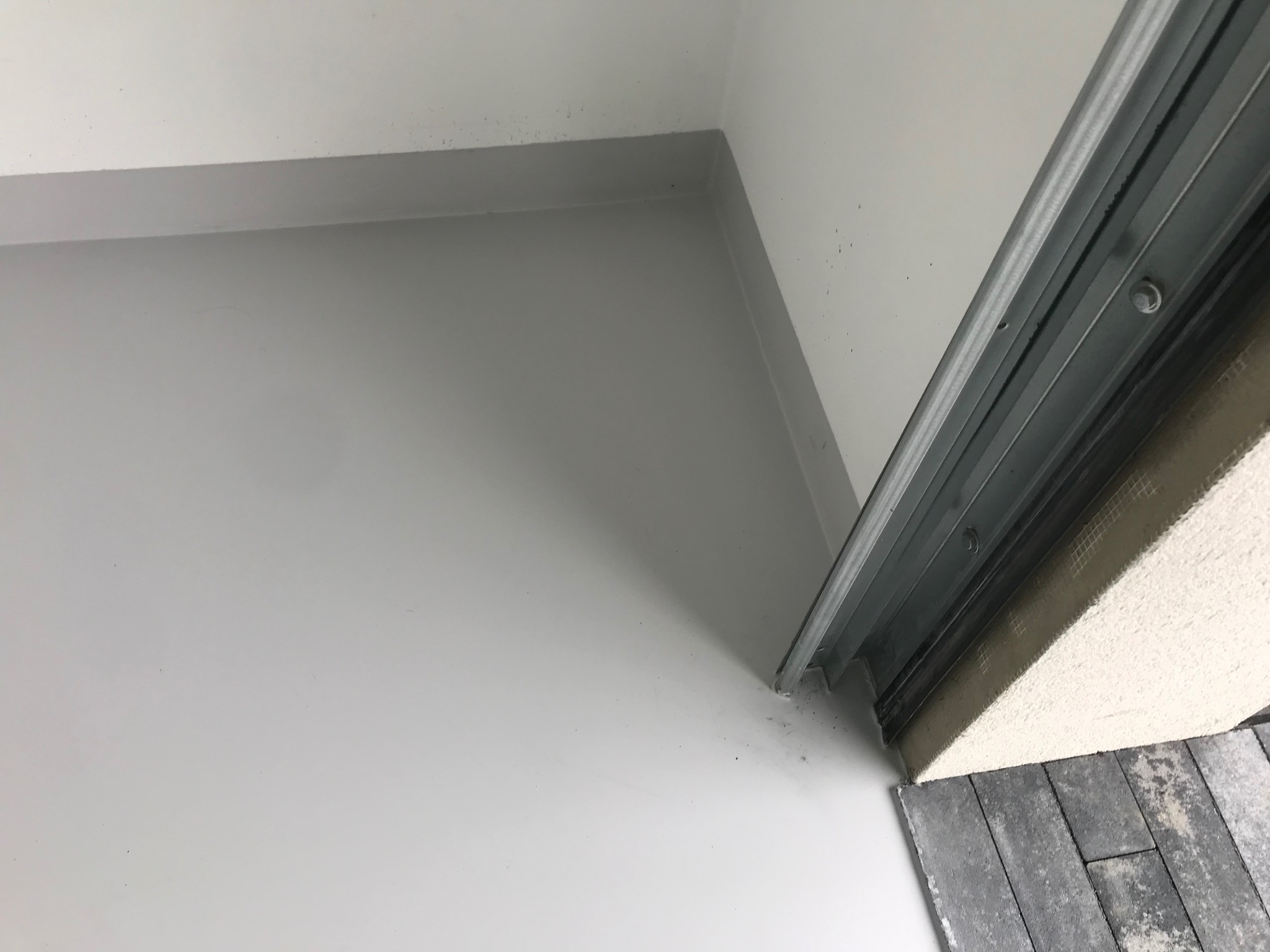 Gładka, jasnoszara powierzchnia podłogi i ściany w rogu pomieszczenia, z widocznym fragmentem szyny i kostki brukowej przy progu. Minimalistyczne wykończenie wnętrza.