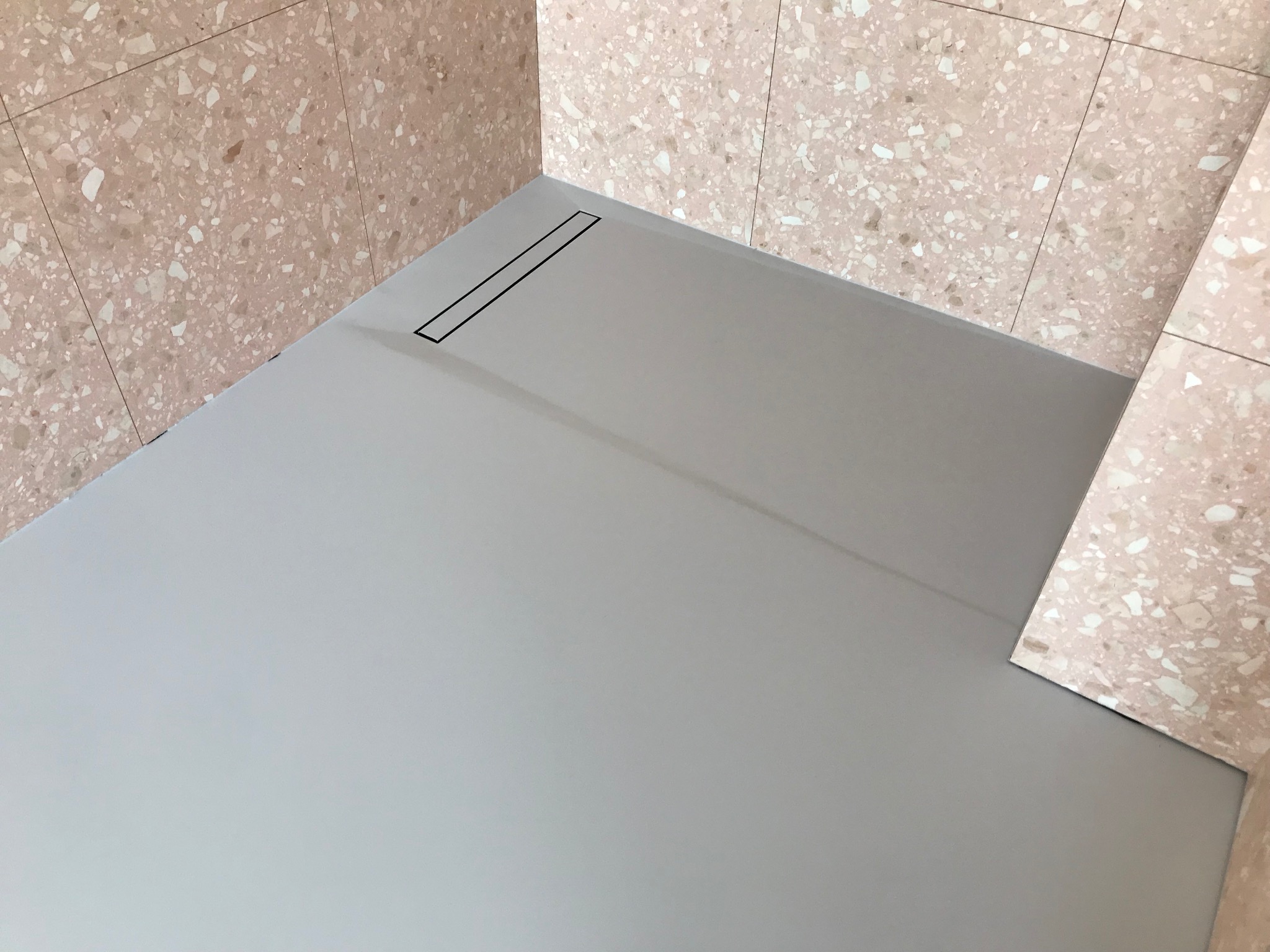 Szara posadzka żywiczna w łazience z odpływem liniowym i ścianami wyłożonymi płytkami imitującymi lastryko w odcieniach różu i beżu. Minimalistyczny design.