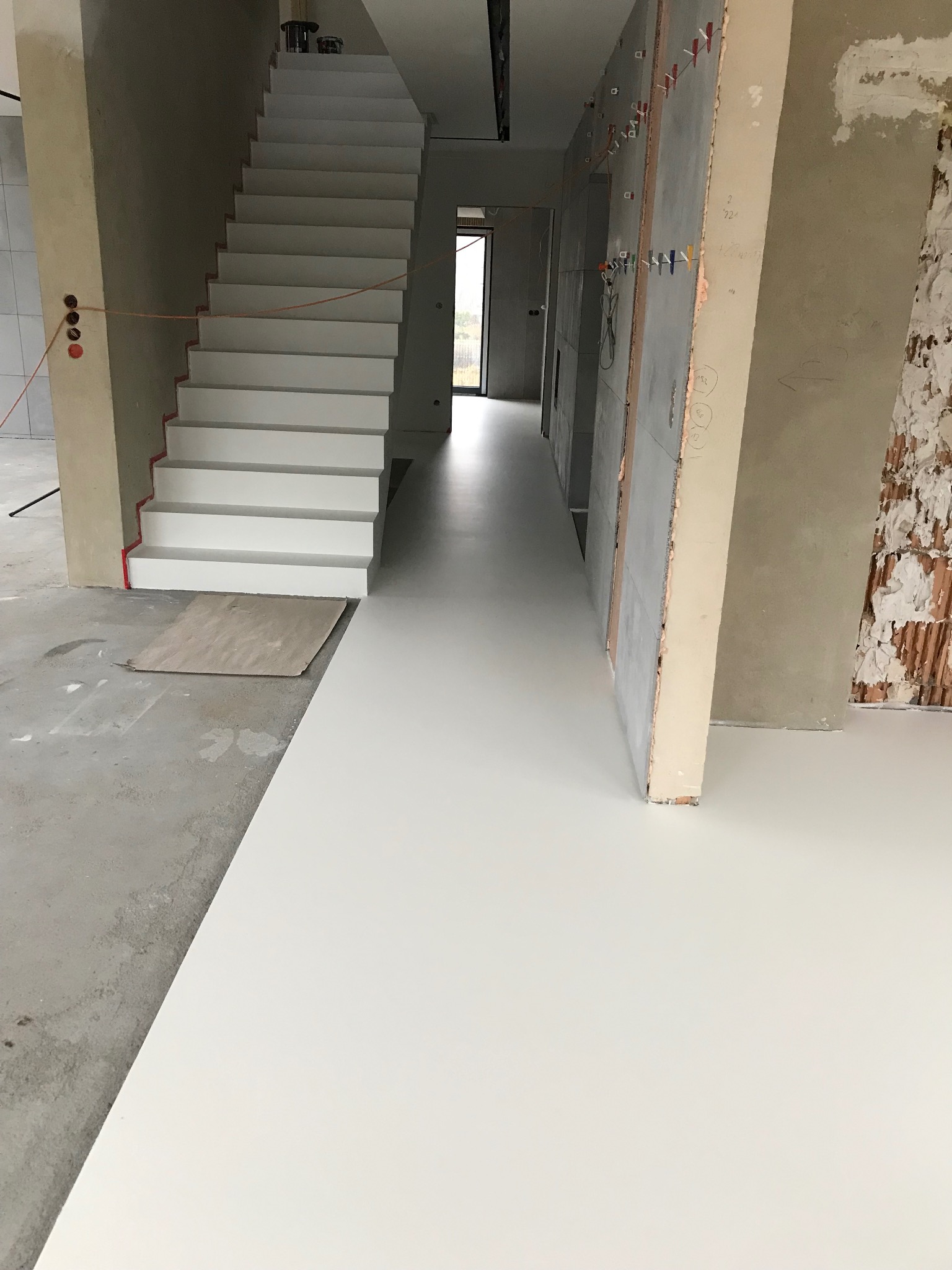 Biała, gładka posadzka w korytarzu z białymi schodami i surowymi ścianami. Widoczne elementy instalacji elektrycznej i fragmenty niezabezpieczonej podłogi.