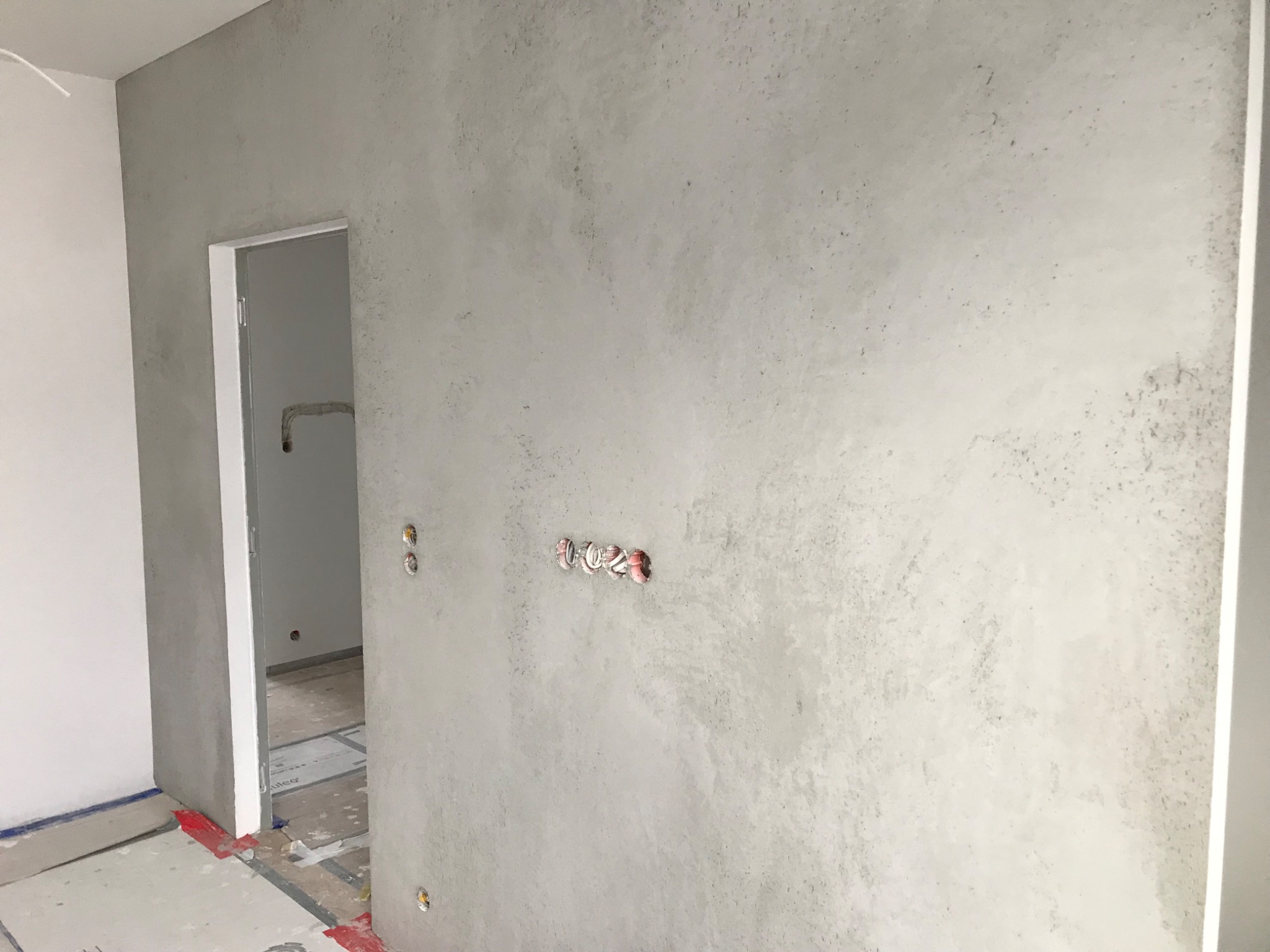 Szara ściana z efektem betonu w nowym wnętrzu, widoczne puszki elektryczne i fragment otwartych drzwi. Minimalistyczny design z surowym wykończeniem.