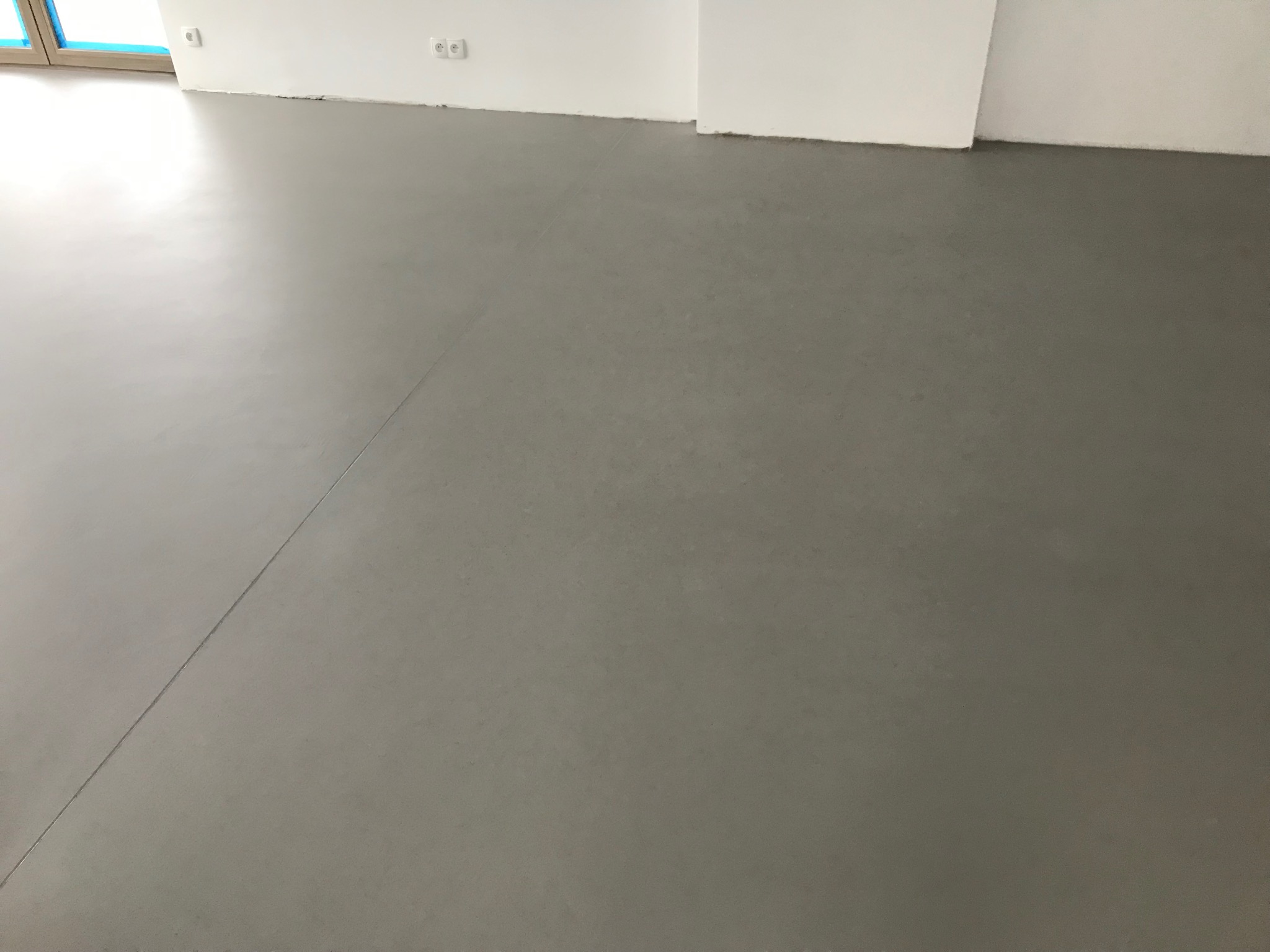 Gładka, szara posadzka w jasnym wnętrzu. Widoczne gniazdka elektryczne na białej ścianie i fragment okna. Minimalistyczny design, jednolite wykończenie podłogi.