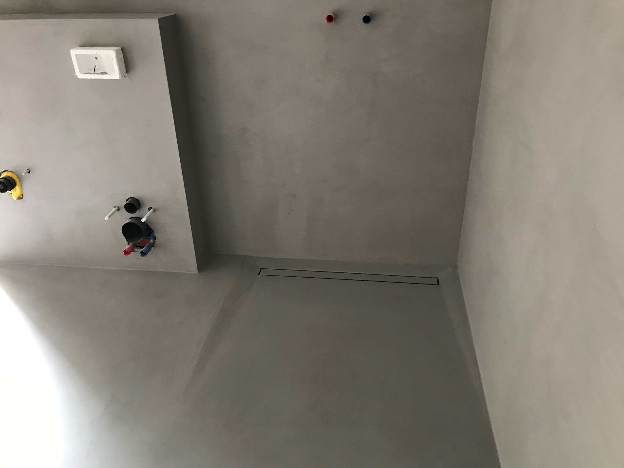 Szara, jednolita powierzchnia podłogi i ścian w łazience z odpływem liniowym i instalacją sanitarną. Minimalistyczne wykończenie wnętrza, surowy styl.