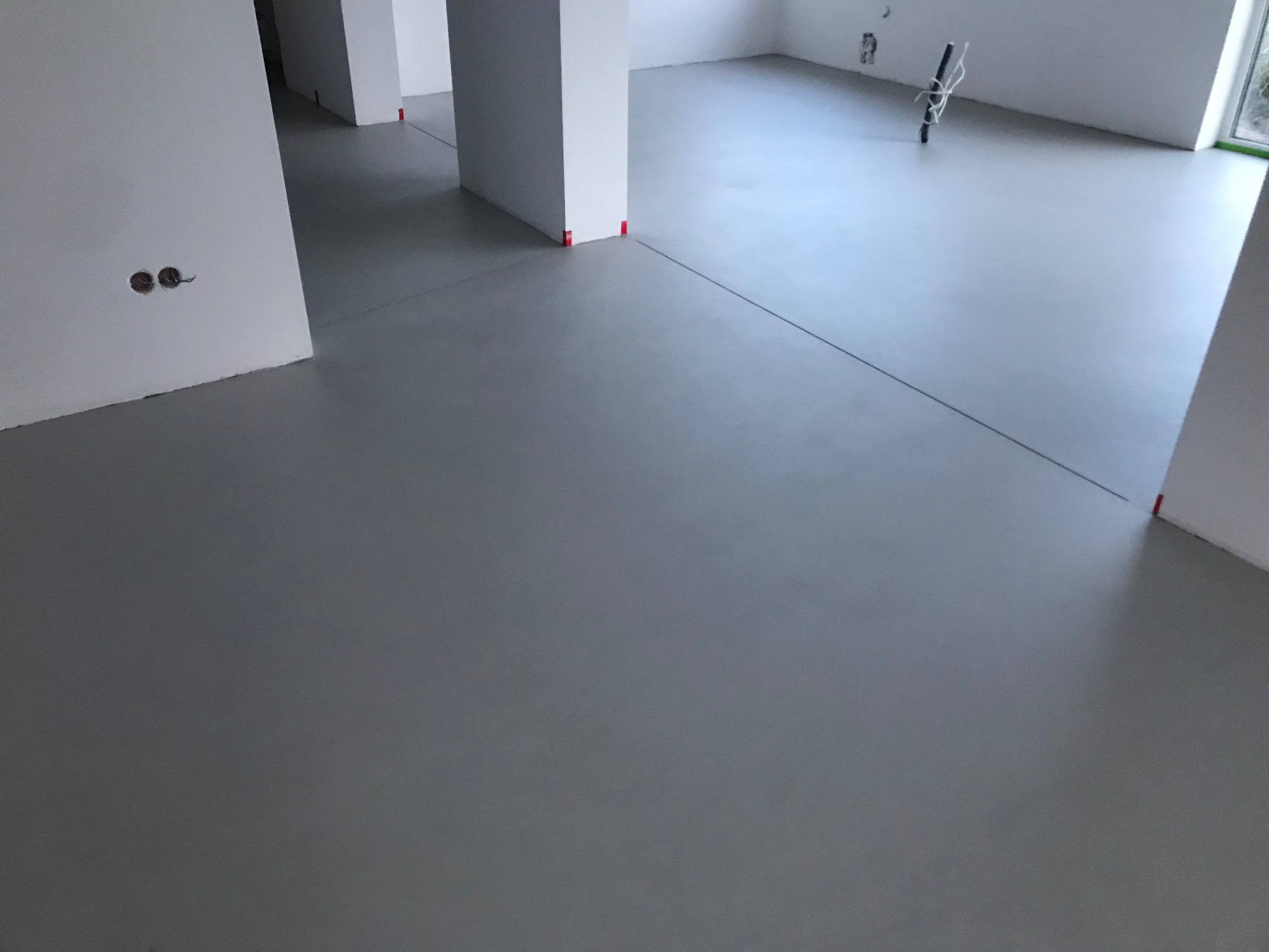Szara, gładka posadzka w jasnym wnętrzu z białymi filarami. Widoczne czerwone znaczniki na filarach i ścianach, oraz wystające kable ze ściany. Minimalistyczny design.
