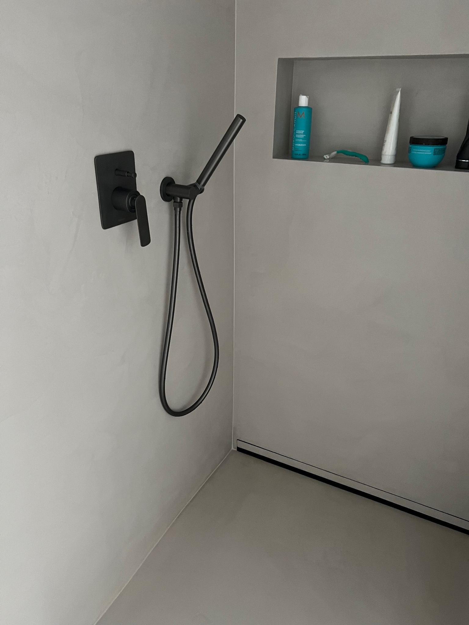 Minimalistyczna łazienka z szarą, gładką powierzchnią ścian i podłogi, czarną armaturą prysznicową i wnęką z kosmetykami. Nowoczesny design.