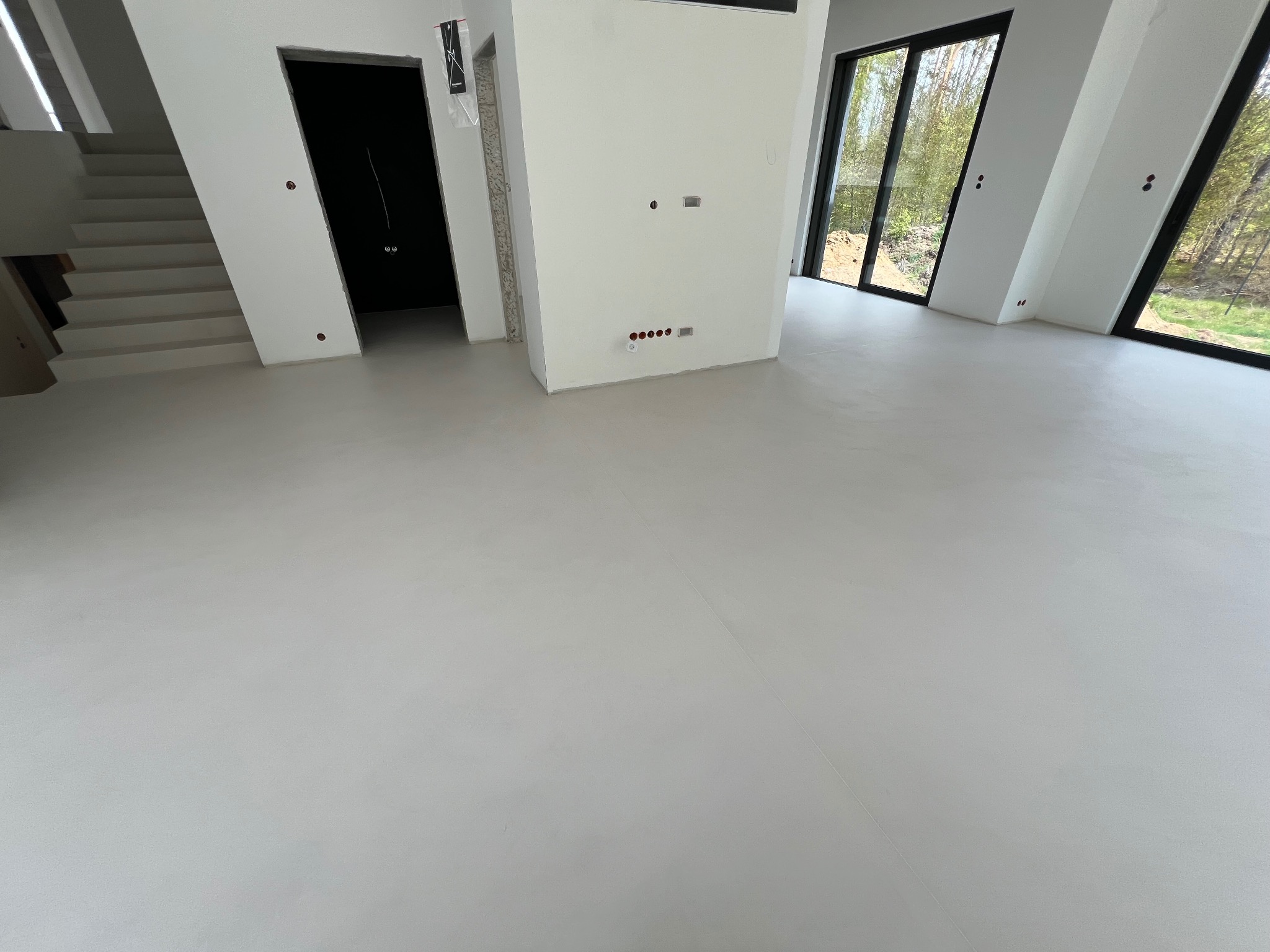 Jasnoszara, gładka posadzka żywiczna w nowoczesnym wnętrzu z dużymi oknami i schodami. Widoczne przygotowane punkty elektryczne na ścianie.