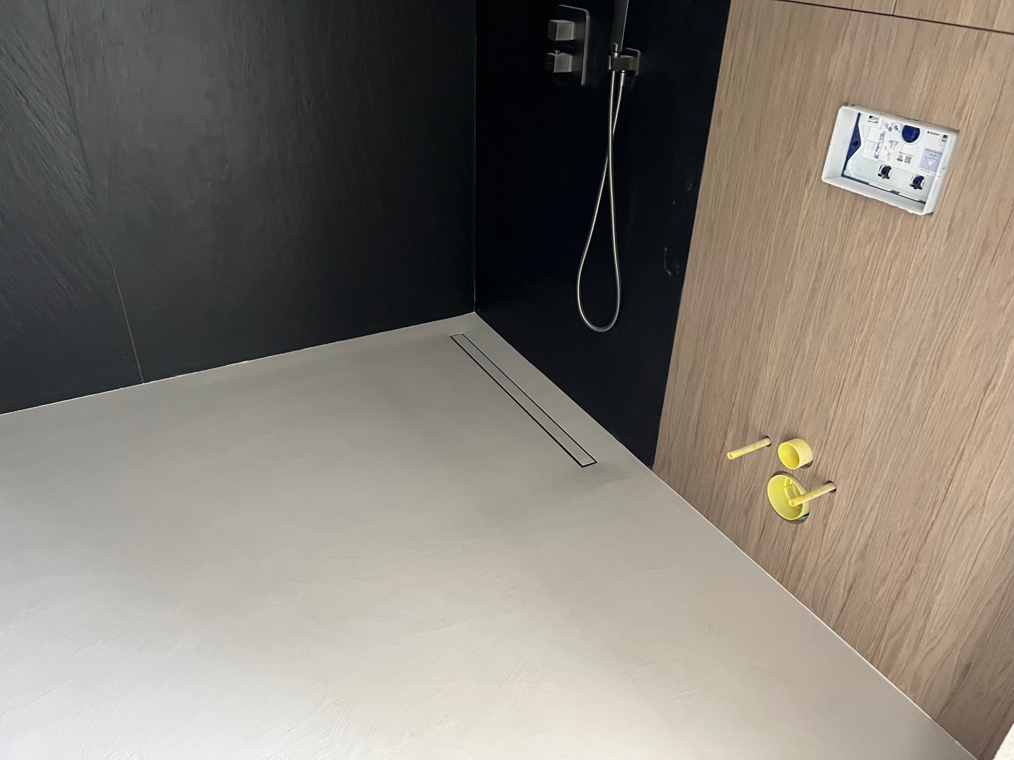 Nowoczesna kabina prysznicowa z szarą, gładką powierzchnią podłogi i czarną ścianą. Widoczny odpływ liniowy, bateria prysznicowa i drewnopodobna obudowa.