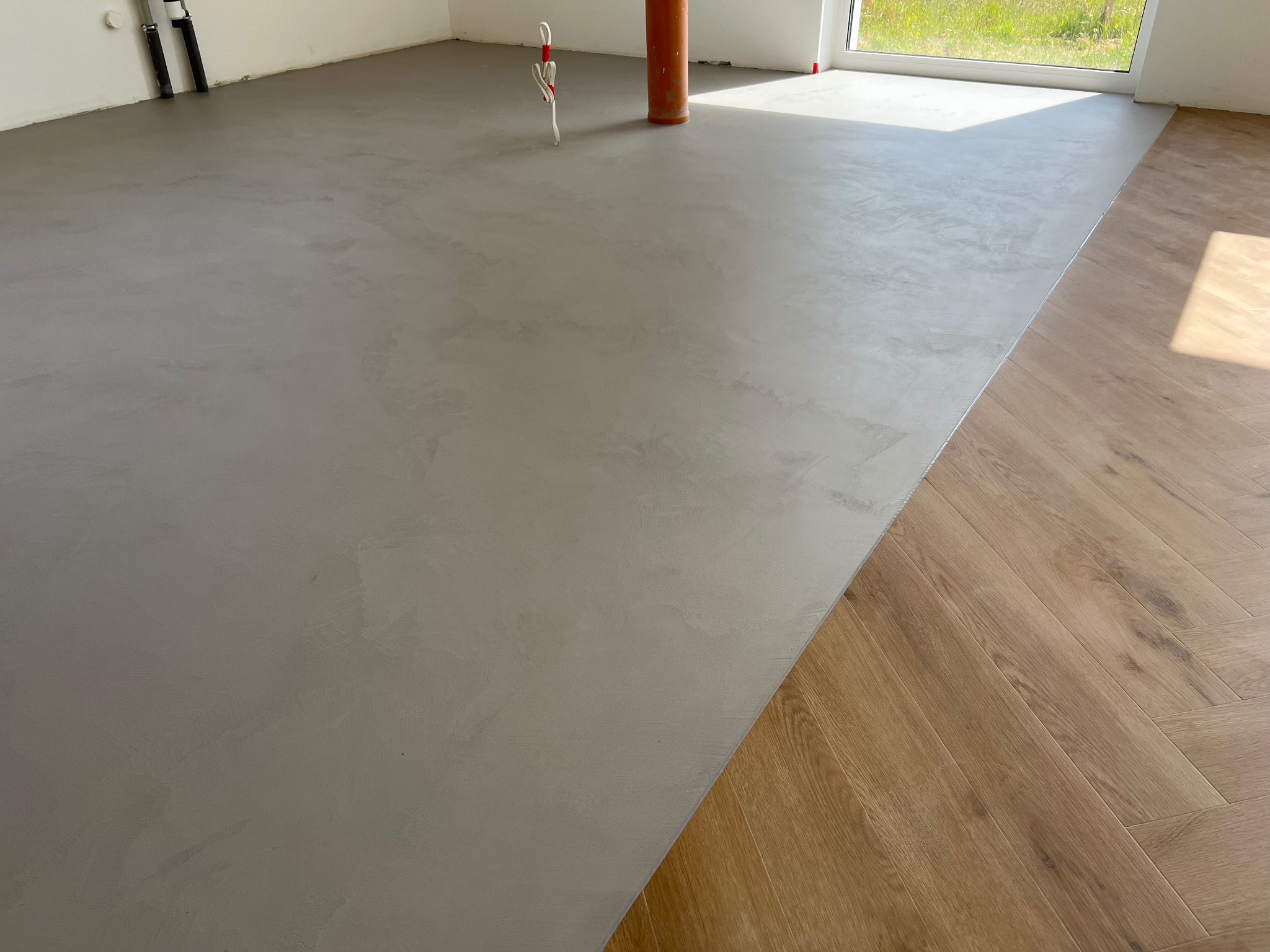 Szara, gładka powierzchnia wylewki podłogowej, częściowo zakryta parkietem ułożonym w jodełkę. Widoczne pionowe rury instalacyjne i fragment okna z zielenią na zewnątrz.