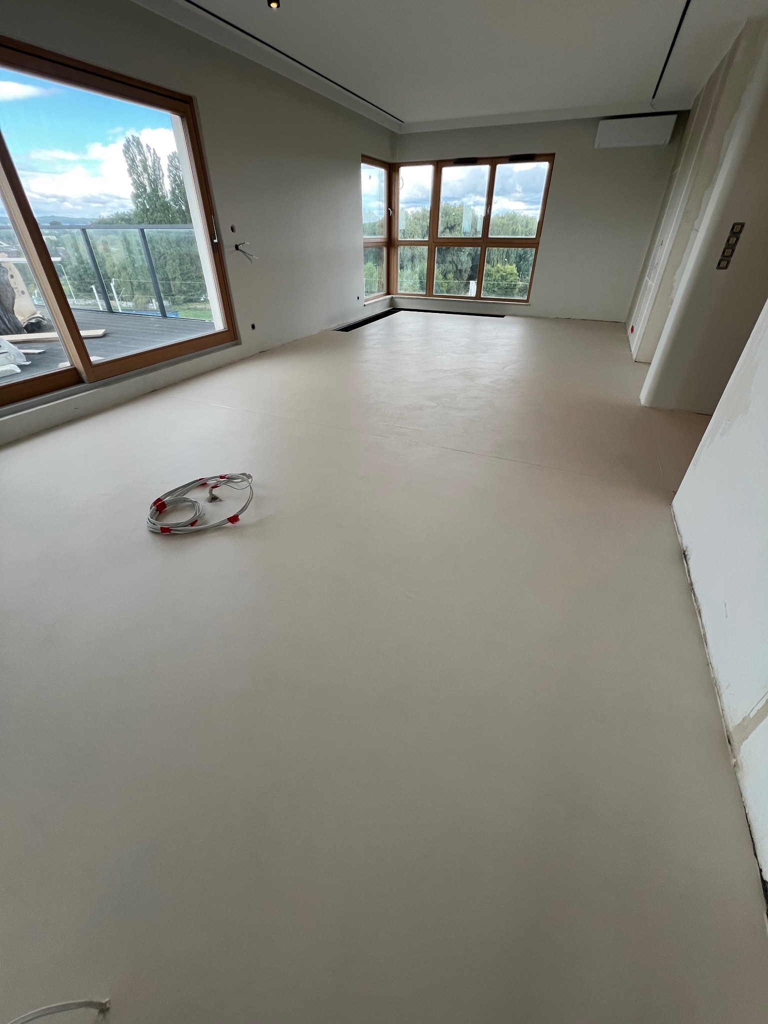 Jasna, gładka podłoga w przestronnym wnętrzu z dużymi oknami. Widoczne kable na podłodze sugerują prace wykończeniowe. Minimalistyczny design, neutralna kolorystyka.