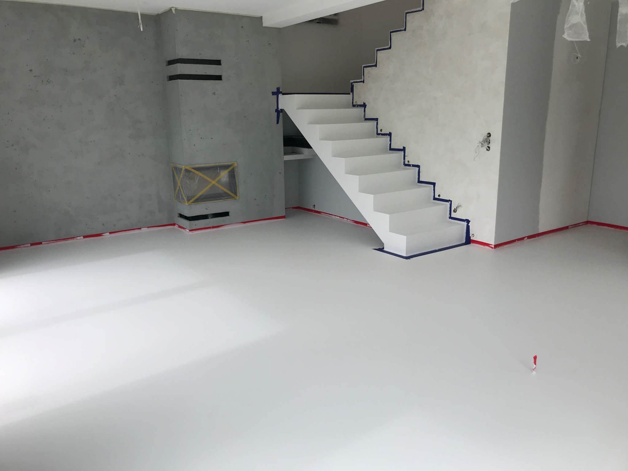 Jasne wnętrze po remoncie: gładka, biała podłoga, szare ściany z fakturą, białe schody z niebieską taśmą zabezpieczającą. Minimalistyczny design, świeżo po realizacji.