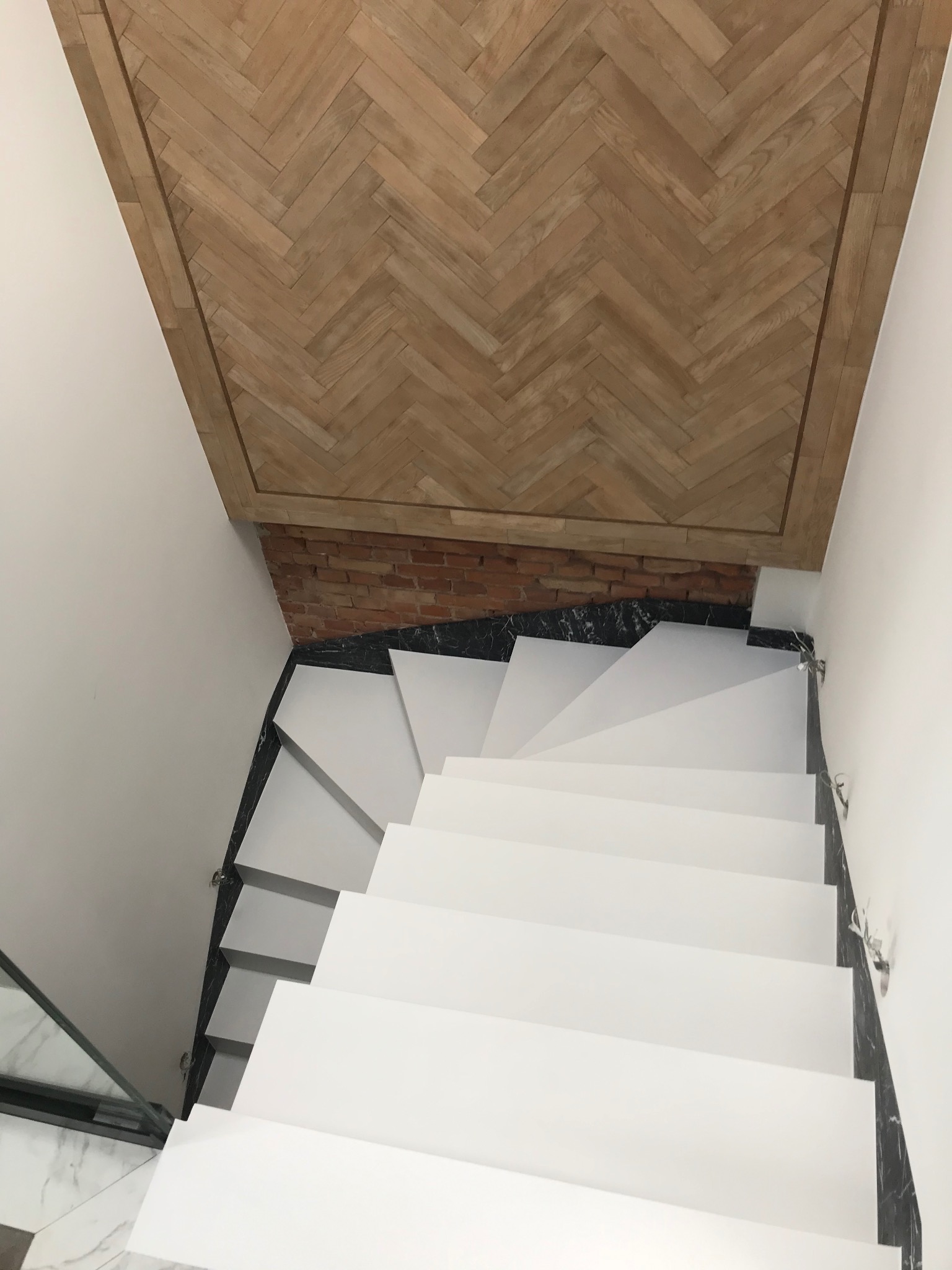 Białe schody z czarnym marmurem u podstawy, drewniany sufit w jodełkę, ceglana ściana. Widok z góry na nowoczesne wykończenie wnętrza.