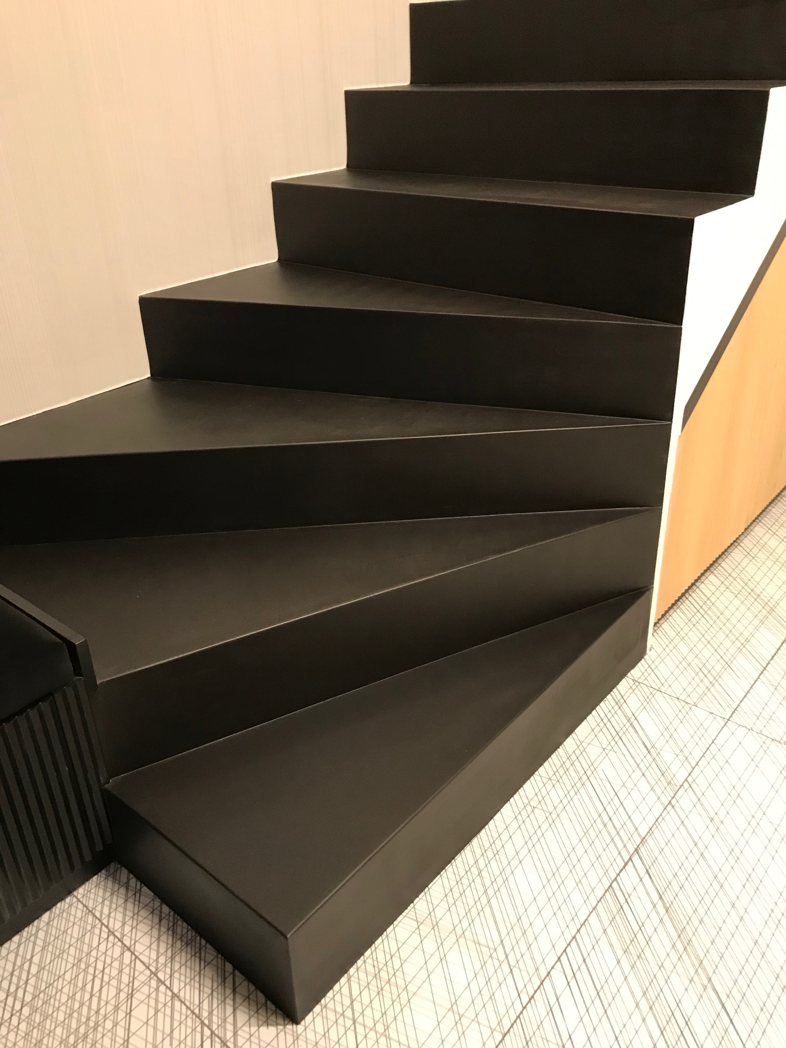 Minimalistyczne, czarne schody z gładką powierzchnią, widoczne z boku, z subtelnym wzorem na podłodze i jasną ścianą w tle. Nowoczesny design.
