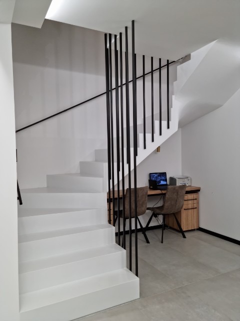 Białe schody z czarną balustradą w nowoczesnym wnętrzu. Widoczny fragment biurka z komputerem i krzesłami. Minimalistyczny design, jasne oświetlenie.