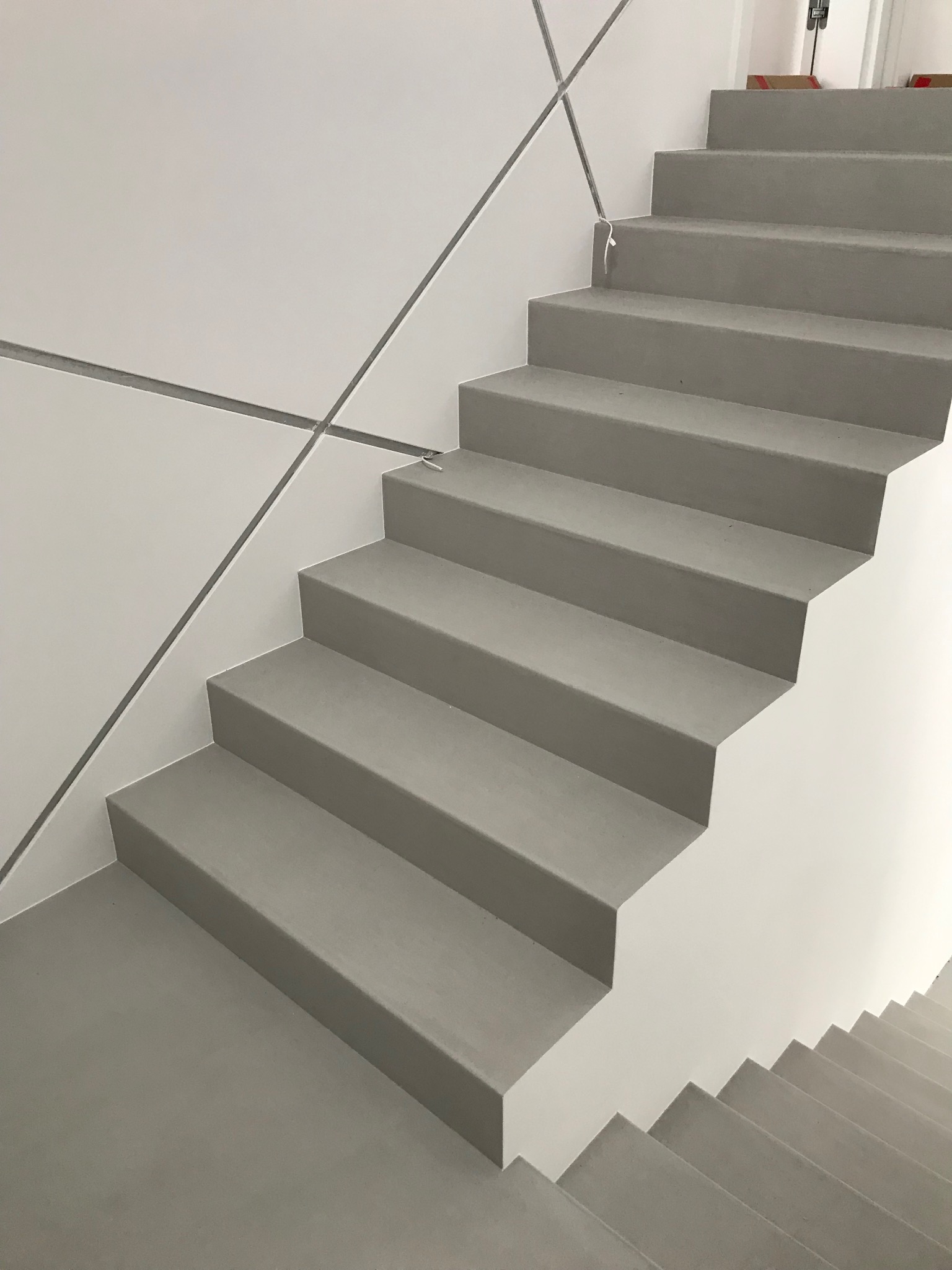 Minimalistyczne schody z szarego materiału, prawdopodobnie beton architektoniczny, z geometrycznymi detalami na ścianie. Widok z dołu, podkreślający prostotę i nowoczesny design.