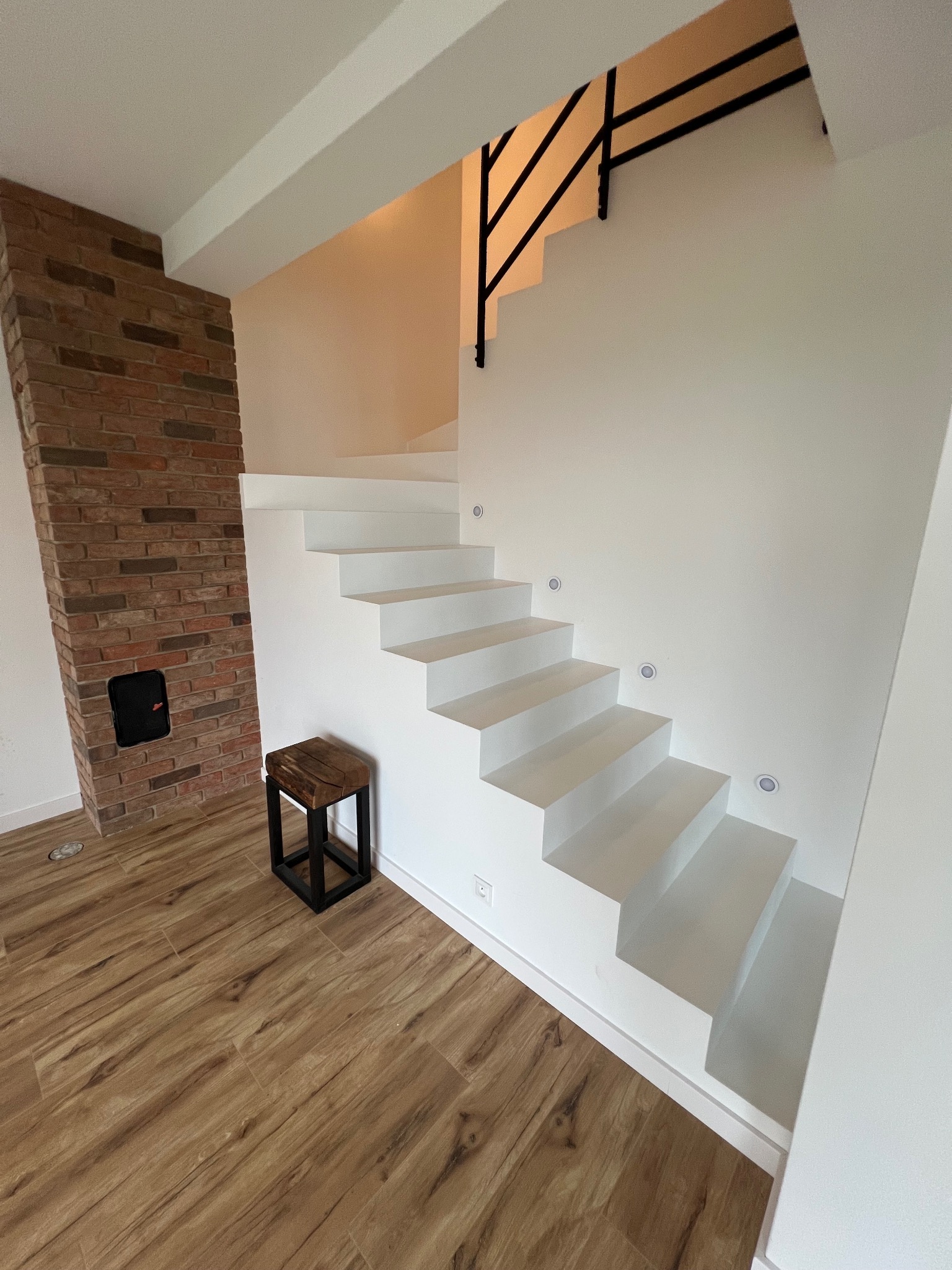 Minimalistyczne, białe schody wspornikowe z czarną balustradą, oświetleniem punktowym i ceglaną ścianą w tle. Podłoga wykończona panelami imitującymi drewno.