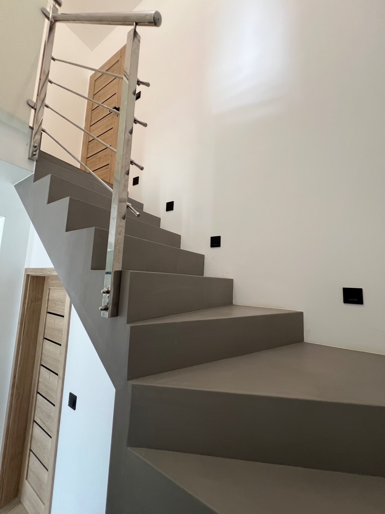 Schody wykończone jednolitą, szarą powierzchnią, z metalową balustradą i minimalistycznymi punktami świetlnymi w ścianie. Widoczne drewniane drzwi w tle.