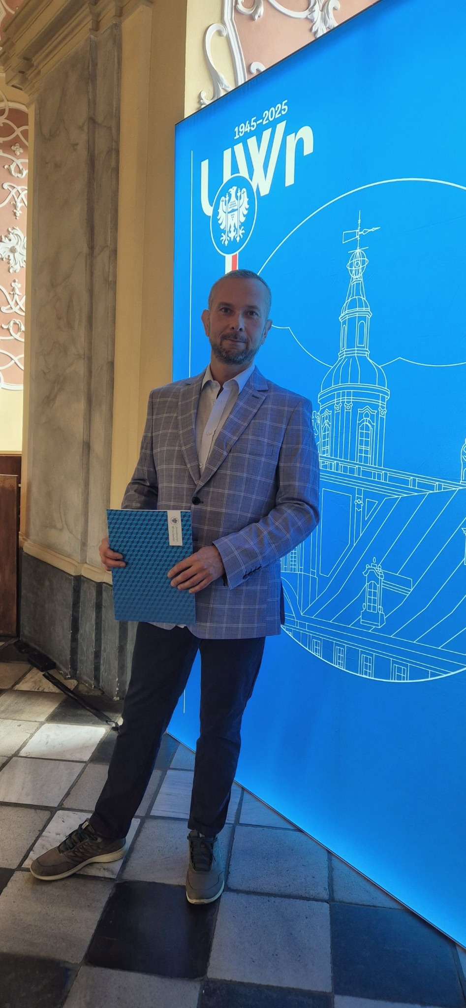 Mężczyzna w marynarce w kratę, trzymający dokument na tle niebieskiej ściany z grafiką Uniwersytetu Wrocławskiego. Uśmiech, kontakt wzrokowy, elegancki strój.